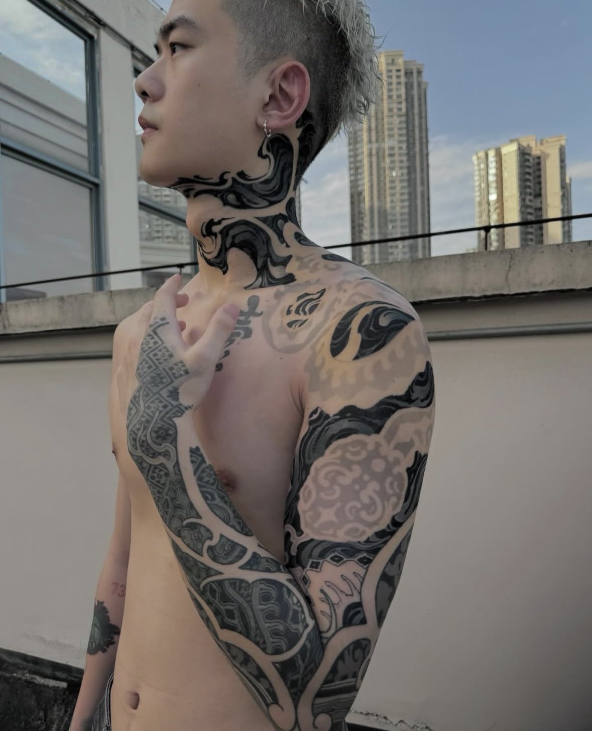 A shirtless young man with short, light-colored hair and extensive black tattoos on his neck, arms, and torso, standing on a rooftop with city buildings in the background.
