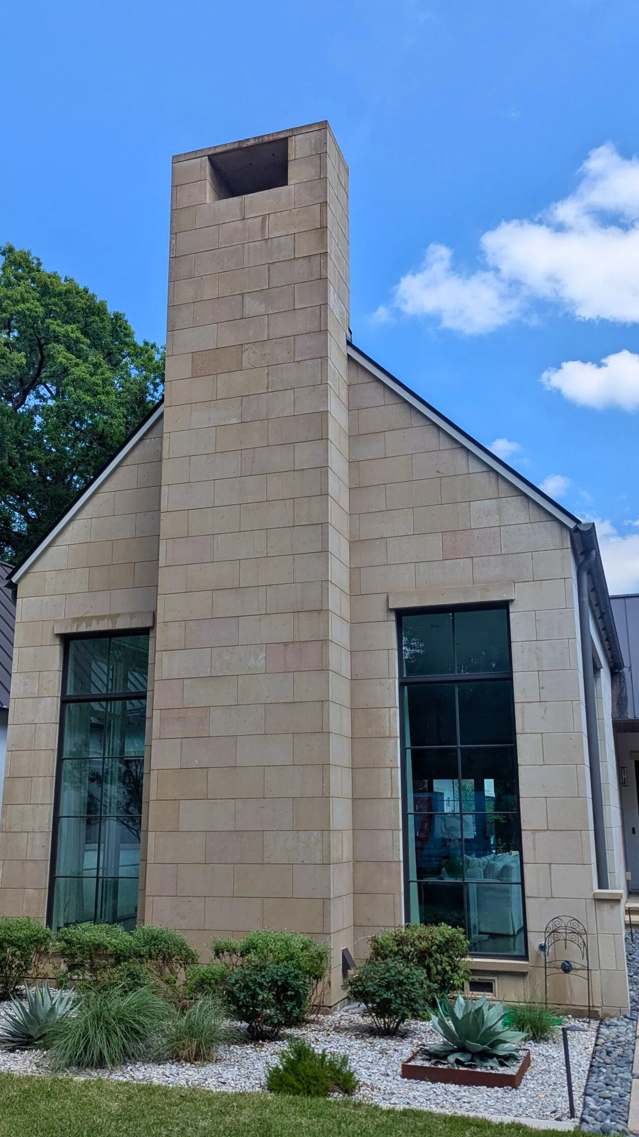 A modern house with a prominent chimney, stone exterior, large windows, and landscaped garden with various plants and shrubs.