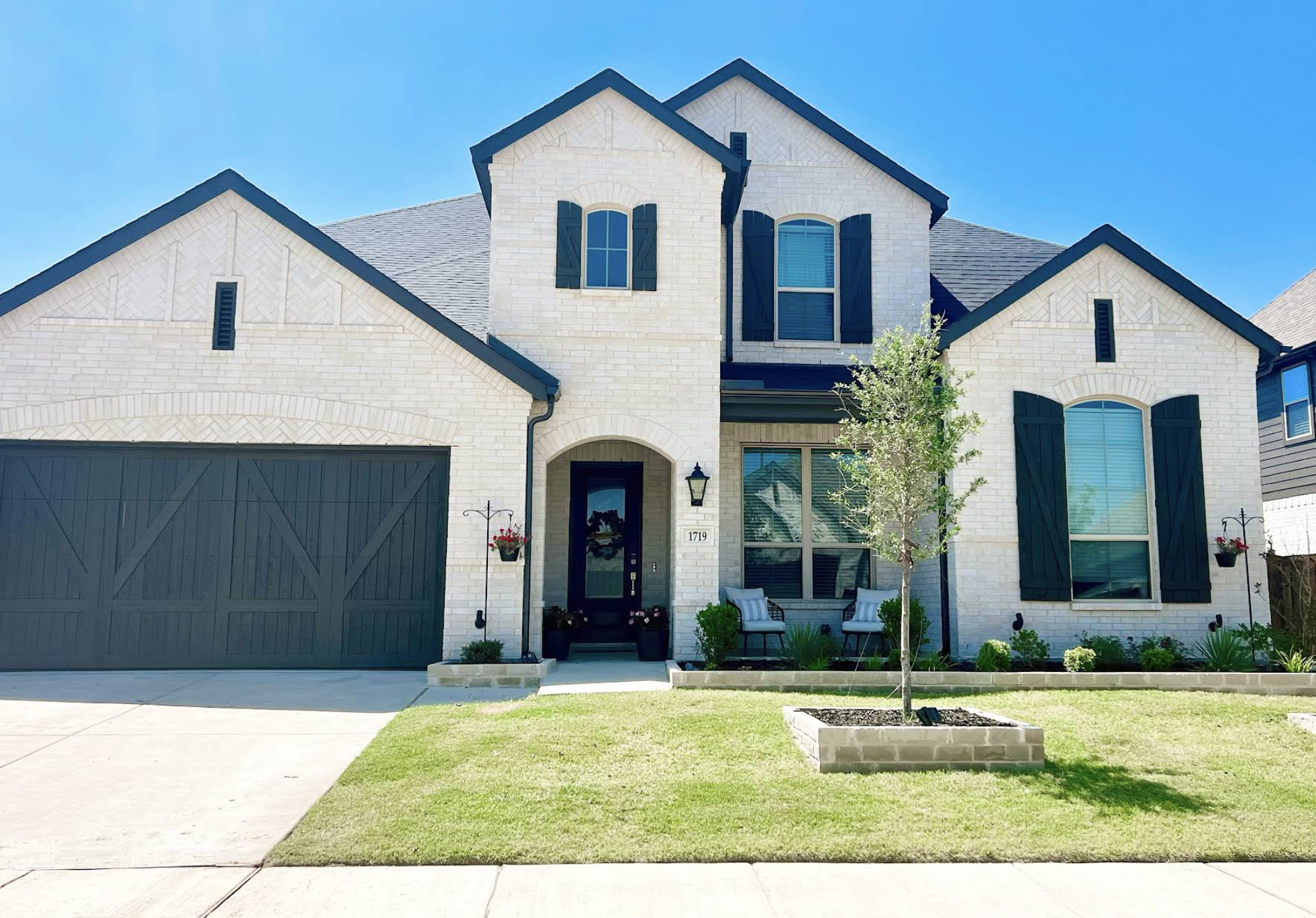 A two-story modern house with white brick exterior, black shutters, and a dark garage door. The front yard has a small lawn, a young tree in a brick-bordered bed, and a seating area with two chairs and a small table.