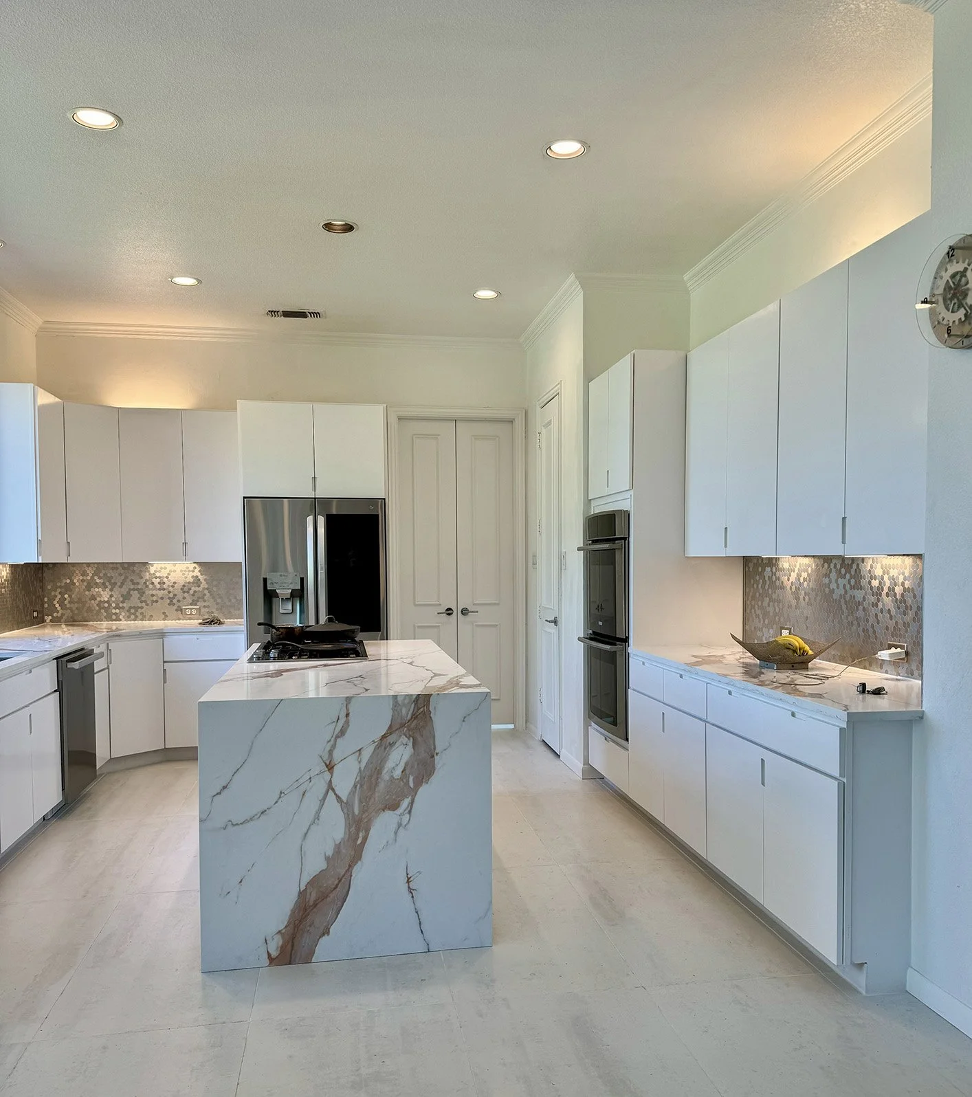 Modern white kitchen with marble island and backsplash, stainless steel refrigerator and oven, white cabinetry, beige floor, ceiling lights, and a wall clock.