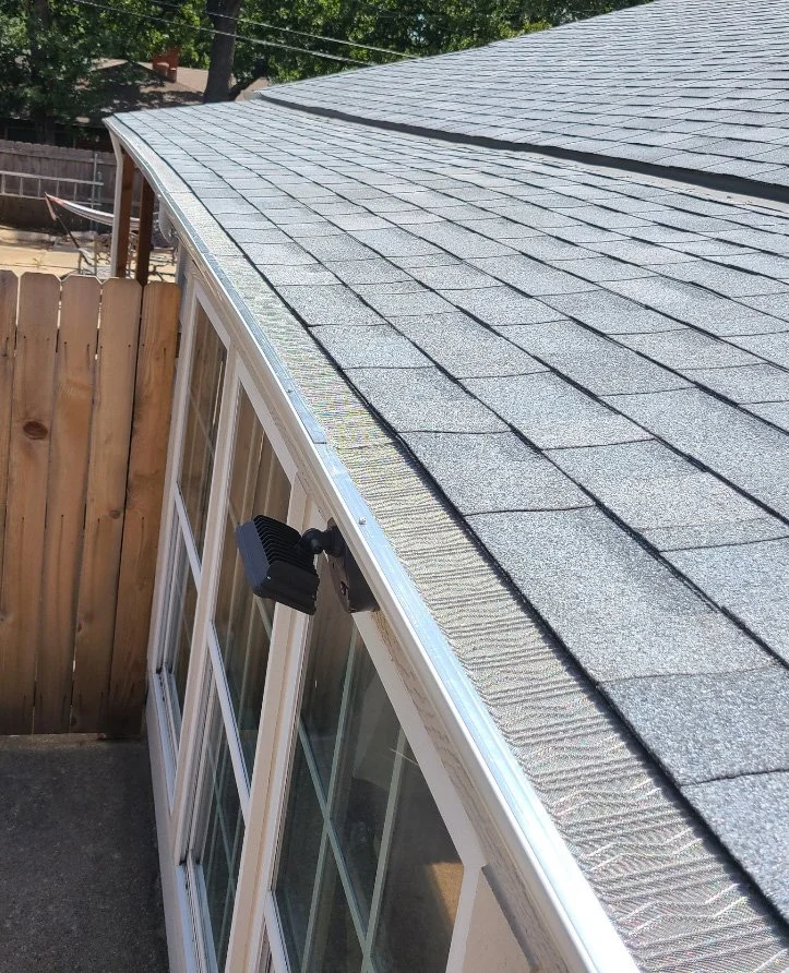 Close-up view of a building's roof with asphalt shingles and a window with security camera mounted nearby, surrounded by a wooden fence.