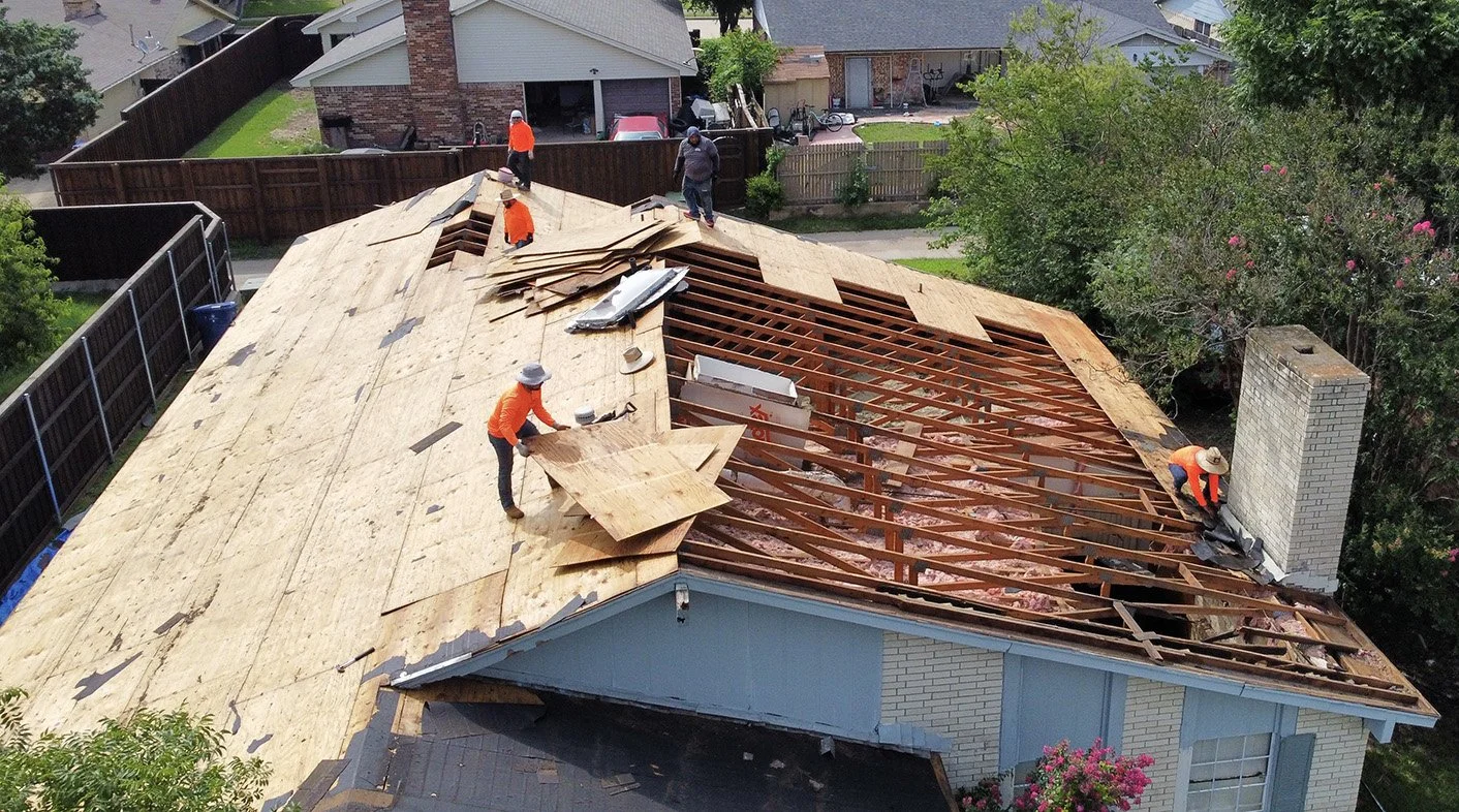 Roof renovation in progress on a house, with workers removing and replacing the shingles, some working on the covered part of the roof, and others on the exposed wooden framework and chimney.