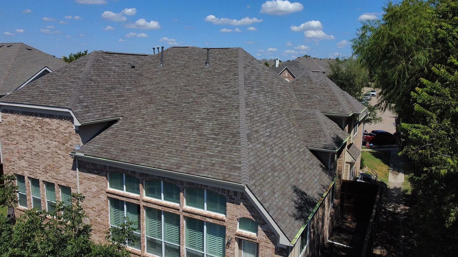 Aerial view of a large brick house with multiple gable roofs covered in gray asphalt shingles, surrounded by trees and a neighborhood with other houses and cars.