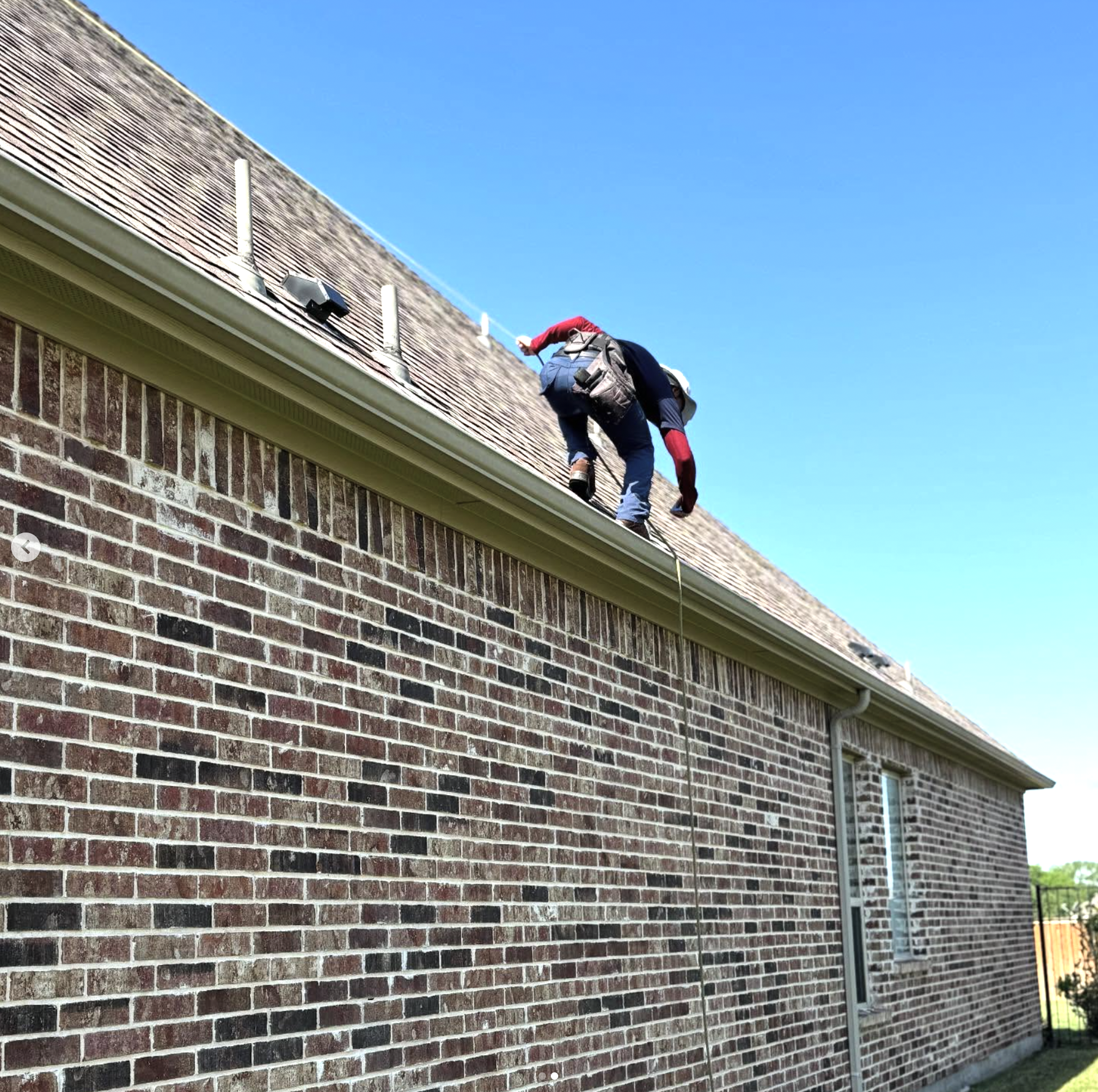 A worker on a ladder cleaning the roof of a brick house during daytime.