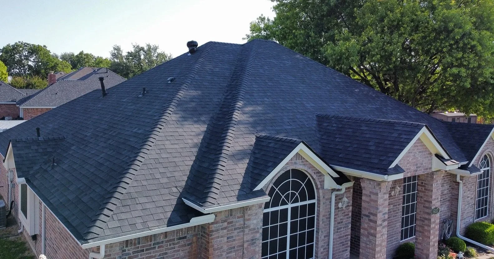 Top view of a brick house with a dark gray shingled roof surrounded by greenery.