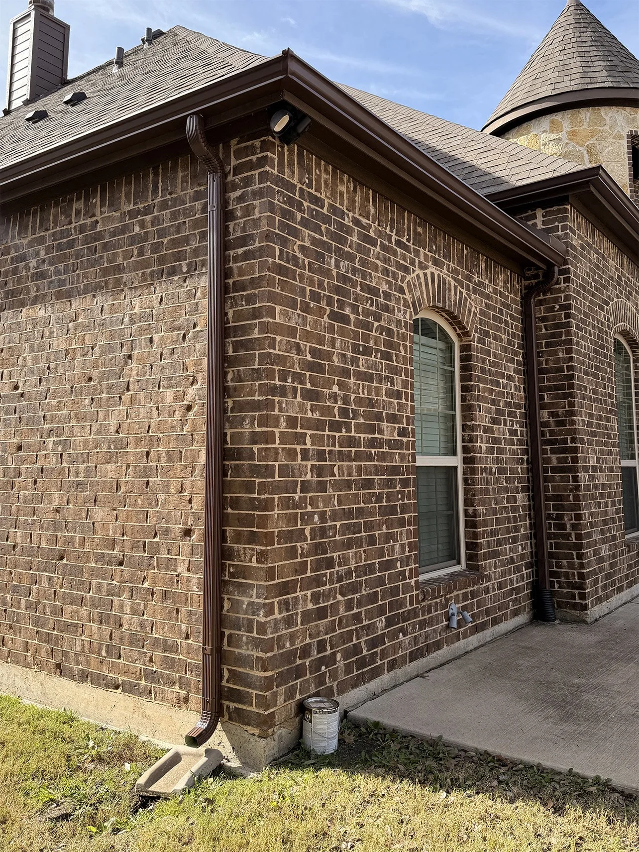 Close-up of a brick house corner with two windows, brown gutters, downspouts, and a small concrete slab at the foundation, under a clear blue sky.