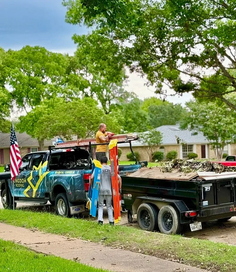Two men working on a black roofing truck with an American flag, parked on a residential street with trees and houses, during daytime.