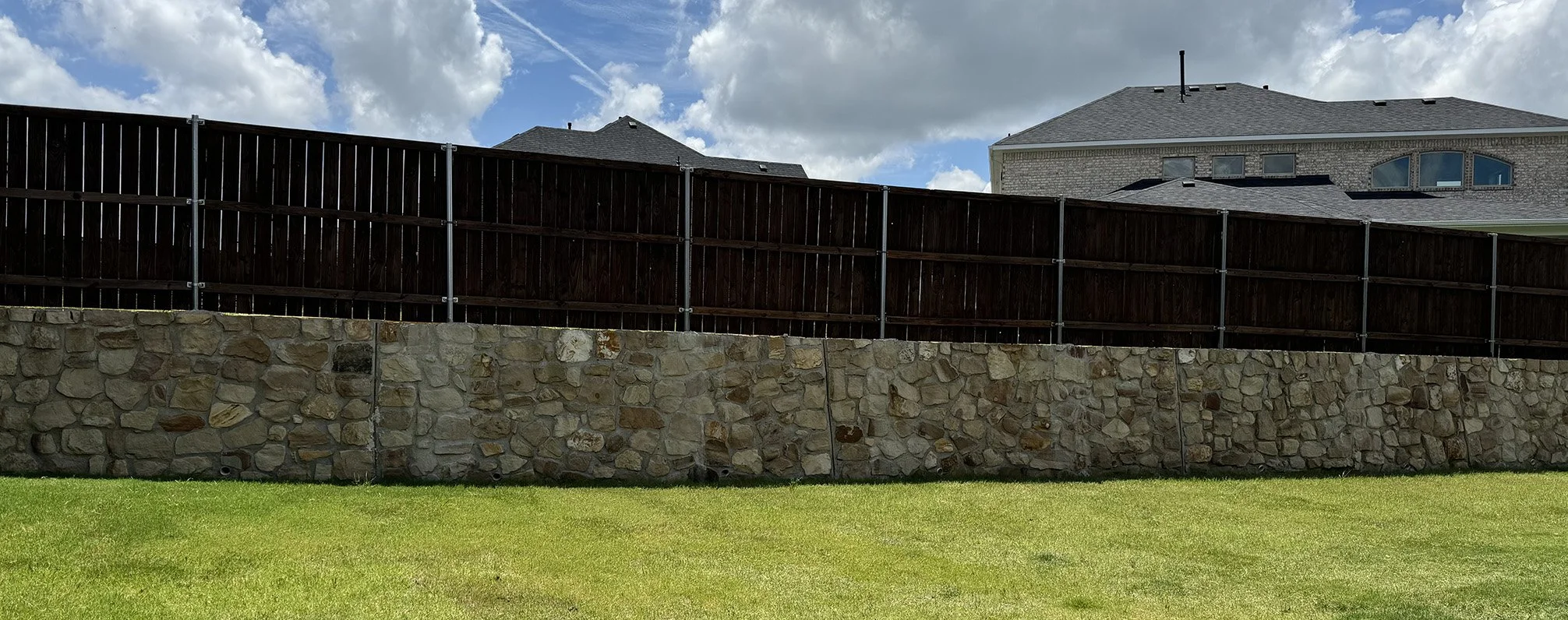 A backyard with a stone retaining wall, a wooden privacy fence, green grass, and a house with a grey roof and multiple windows under a partly cloudy sky.