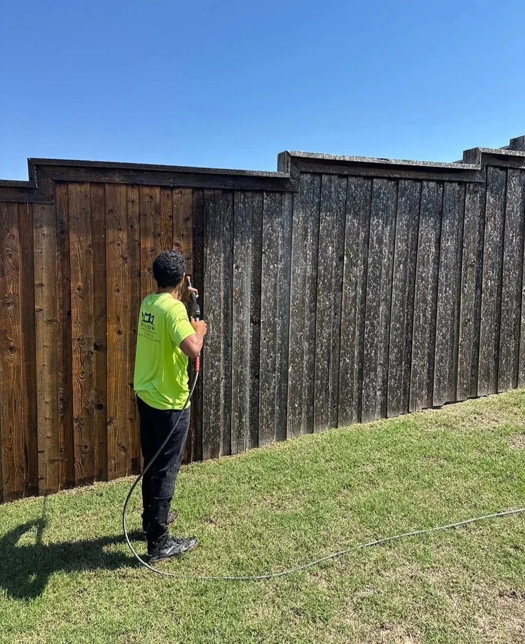 A person cleaning or washing a wooden fence with a pressure washer on a sunny day.