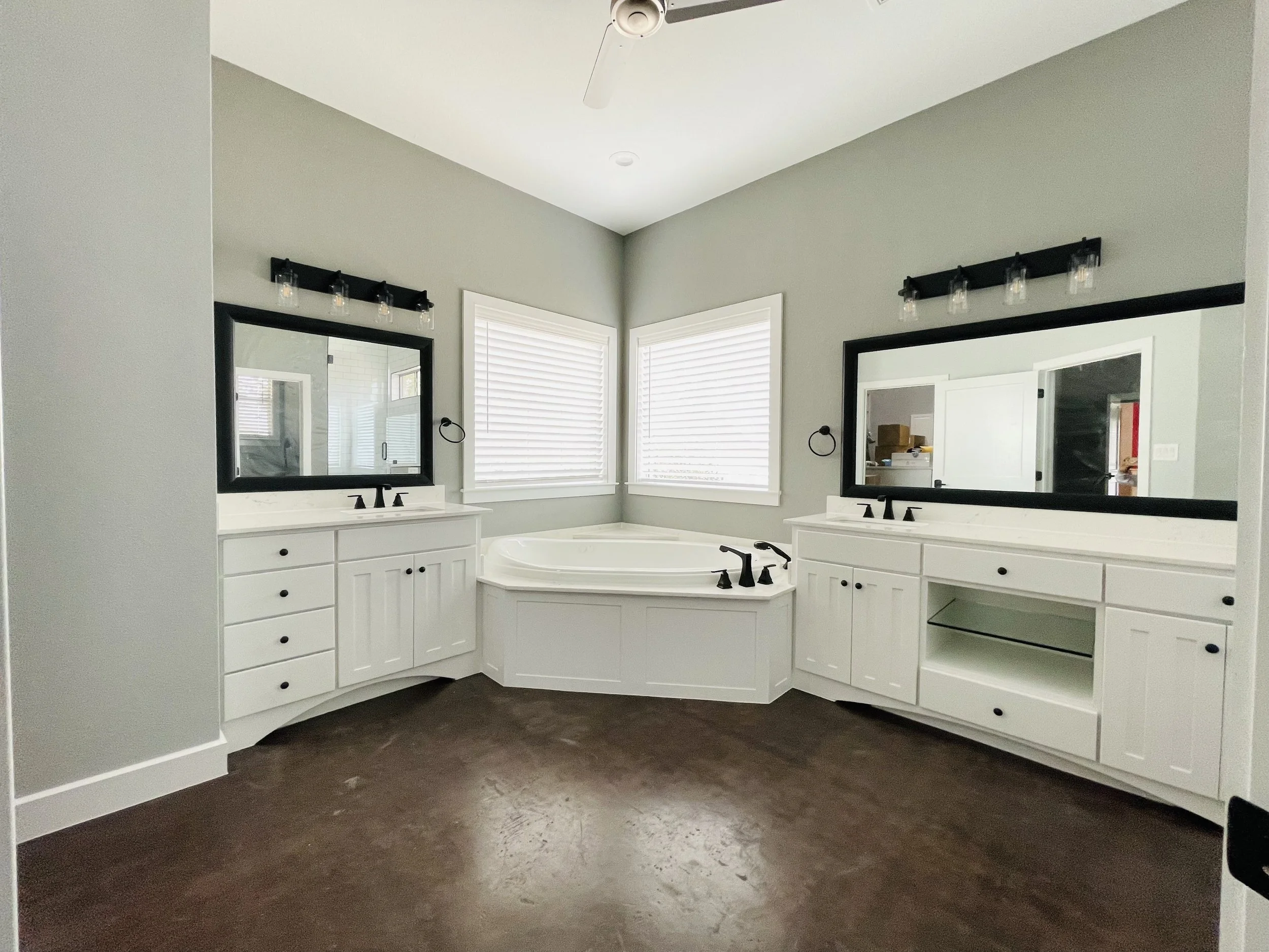A modern bathroom with a corner bathtub, two white vanities with black fixtures, large black-framed mirrors, and wood flooring. Two windows with white blinds provide natural light.