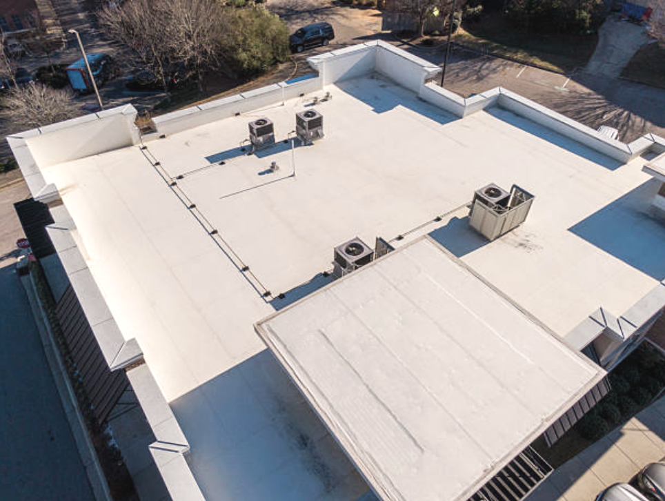 A flat rooftop of a building with three HVAC units and some structural features, surrounded by trees and a parking lot.