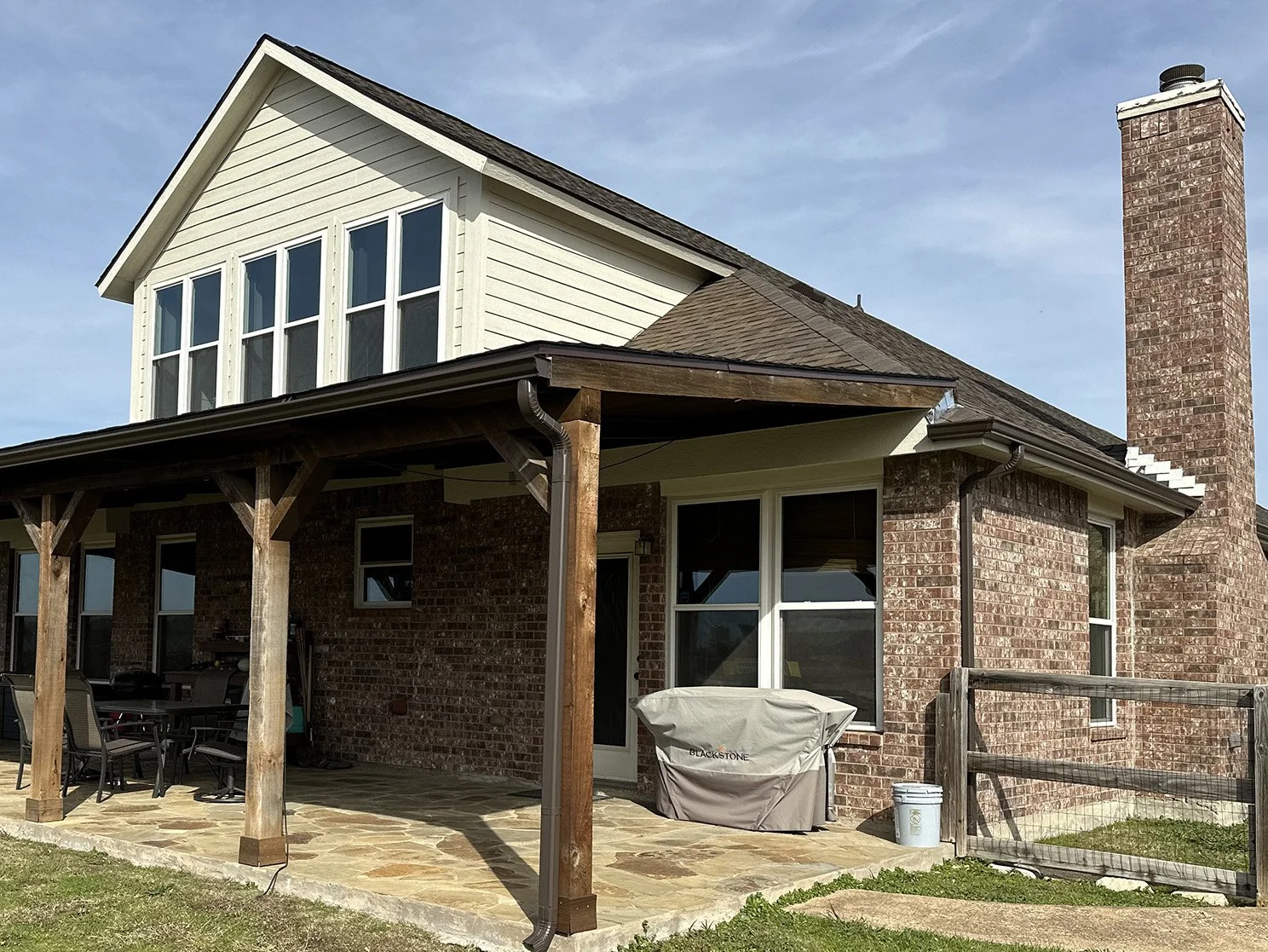 Rear view of a brick house with a wooden porch, patio furniture, a grill covered with a tarp, and a tall brick chimney, under a partly cloudy sky.