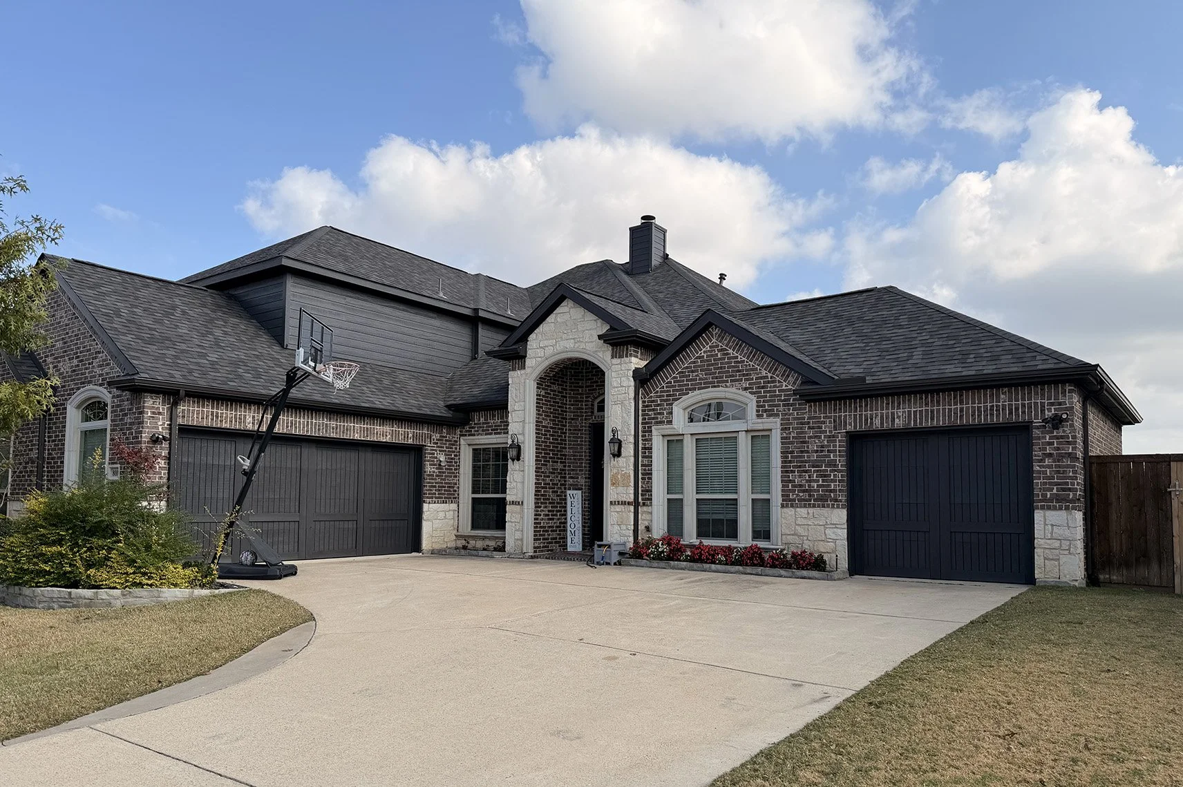 Front view of a modern brick house with two attached dark garage doors, a concrete driveway, and a basketball hoop on a stand. There are windows with white frames, a small flower bed with red flowers, a welcome sign, and a clear blue sky with some clouds.