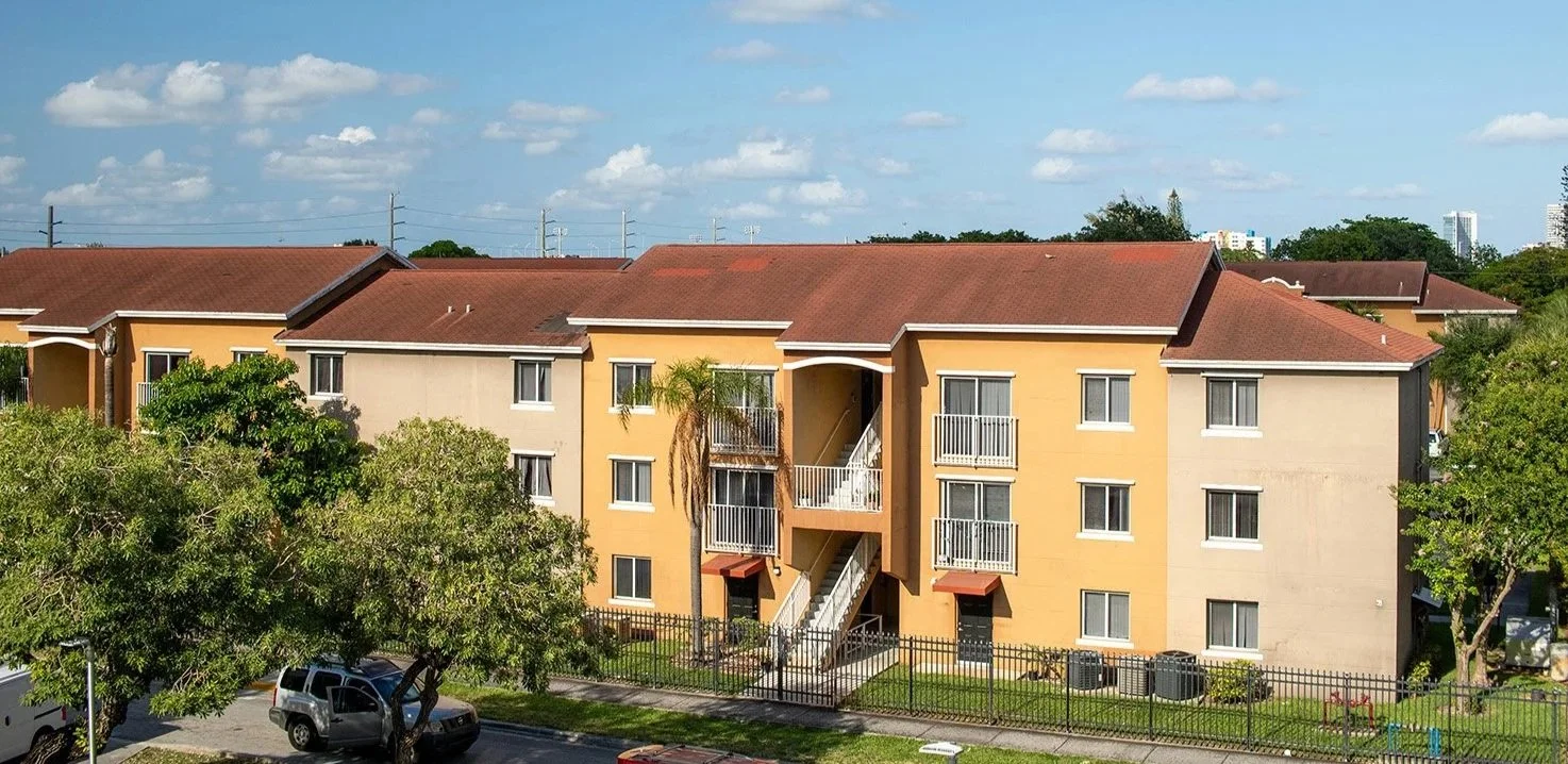 Multi-story apartment building with orange and beige exterior walls, balconies with white railings, and a red-tiled roof, surrounded by trees and a parking lot.