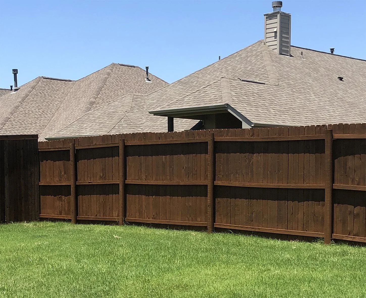 A backyard with a green lawn, wooden fence, and house rooftops with beige shingles and chimneys in the background under a clear blue sky.