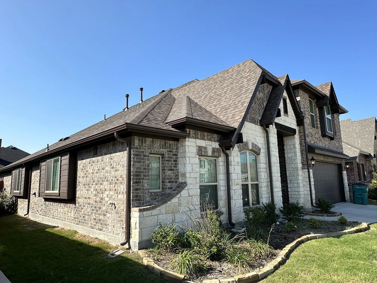 Exterior view of a modern two-story house with brick and stone facade, multiple windows, a grey garage door, and a small garden in front, under a clear blue sky.