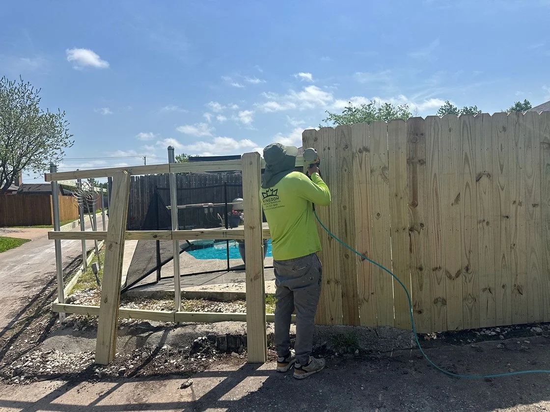 A worker in a yellow shirt and gray pants is installing a wooden fence next to a swimming pool in a backyard on a sunny day.