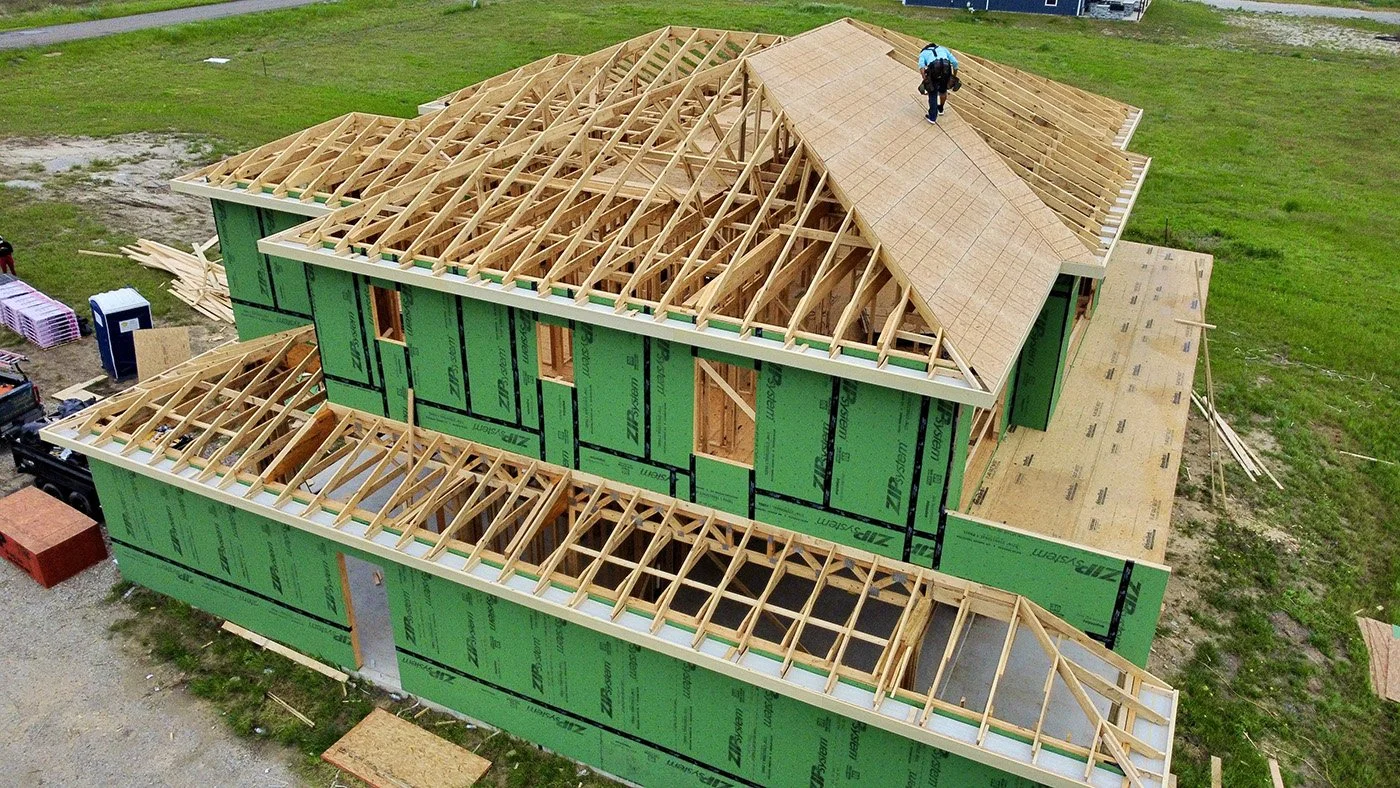 An aerial view of a house under construction showing wooden roof framing and green weather-resistant sheathing. One worker is visible on the roof.