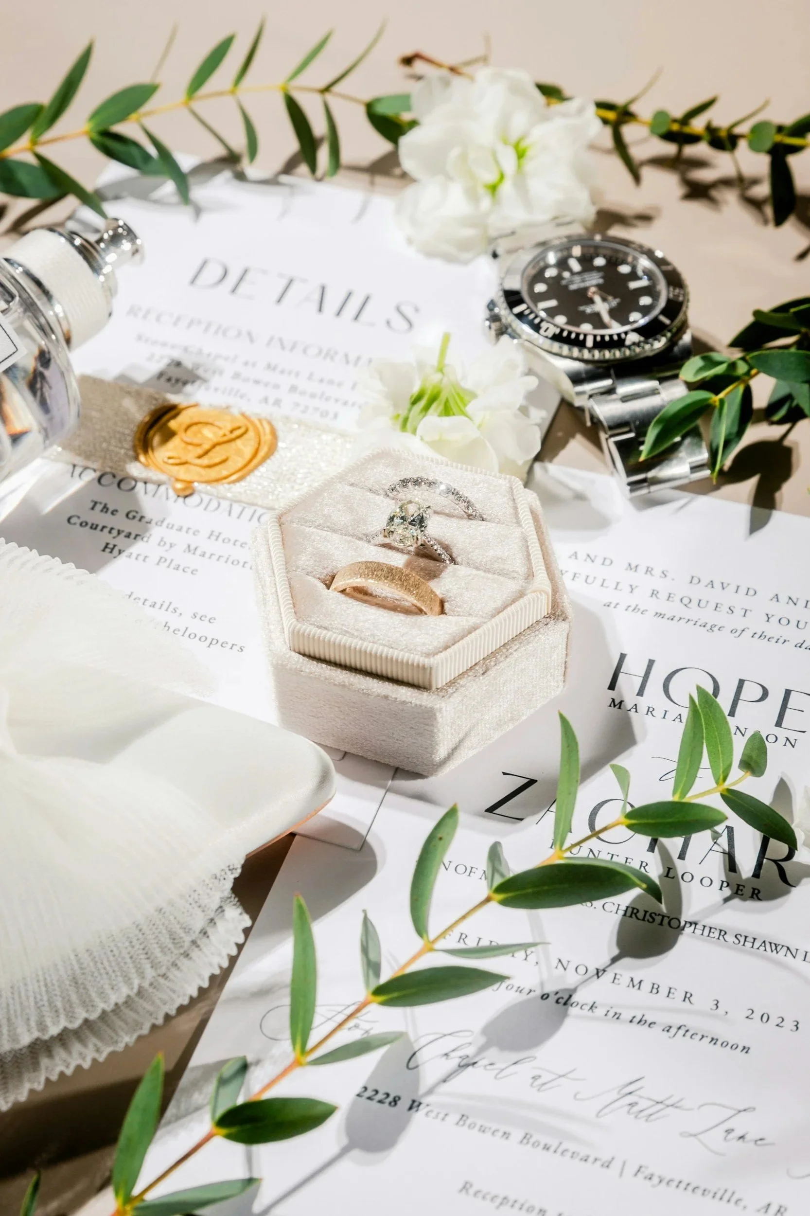 A flat lay of wedding details including a diamond engagement ring in a velvet box, wedding rings, a wedding invitation, greenery, a watch, a perfume bottle, a wax seal, and white flowers.