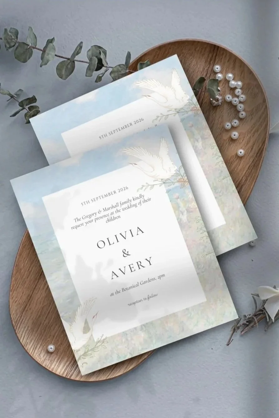 Wedding invitation with a soft watercolor background featuring white cranes, placed on a wooden tray with white pearl beads and greenery nearby.