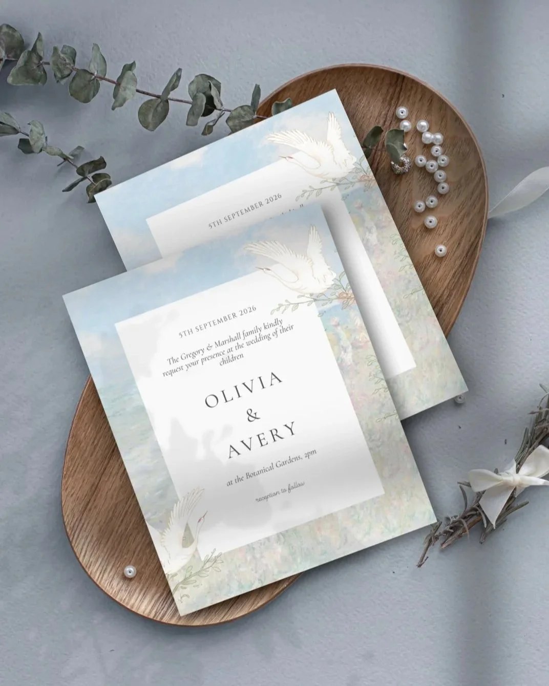 Wedding invitation cards with a nature and bird theme, placed on a wooden tray, accompanied by pearl beads, eucalyptus leaves, and a small bundle of greenery tied with a white ribbon.