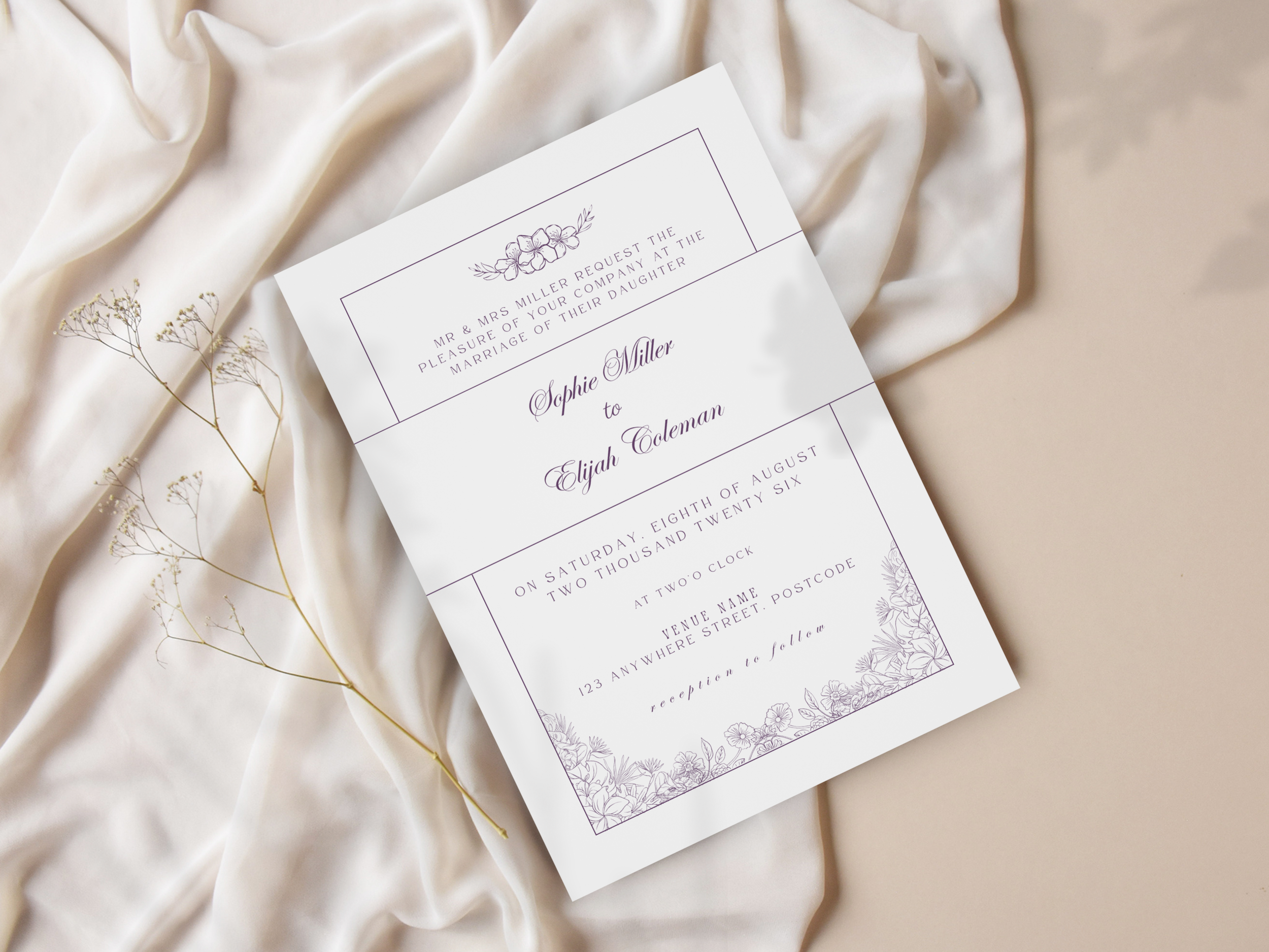 Wedding invitation card with floral design, placed on a soft, cream-colored fabric, with a small sprig of dried flowers beside it.
