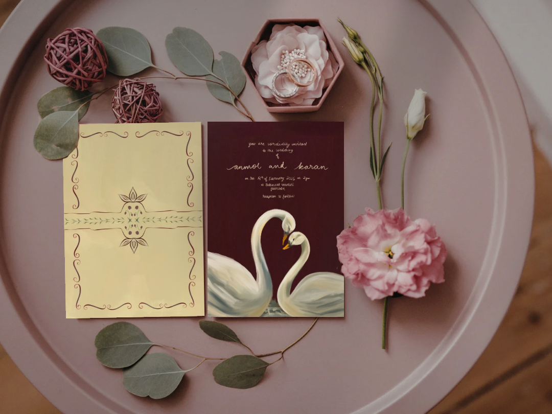 Wedding invitation card featuring two swans forming a heart, surrounded by pink and green decorative elements, with pink and white flowers and green leaves arranged on a pink tray.