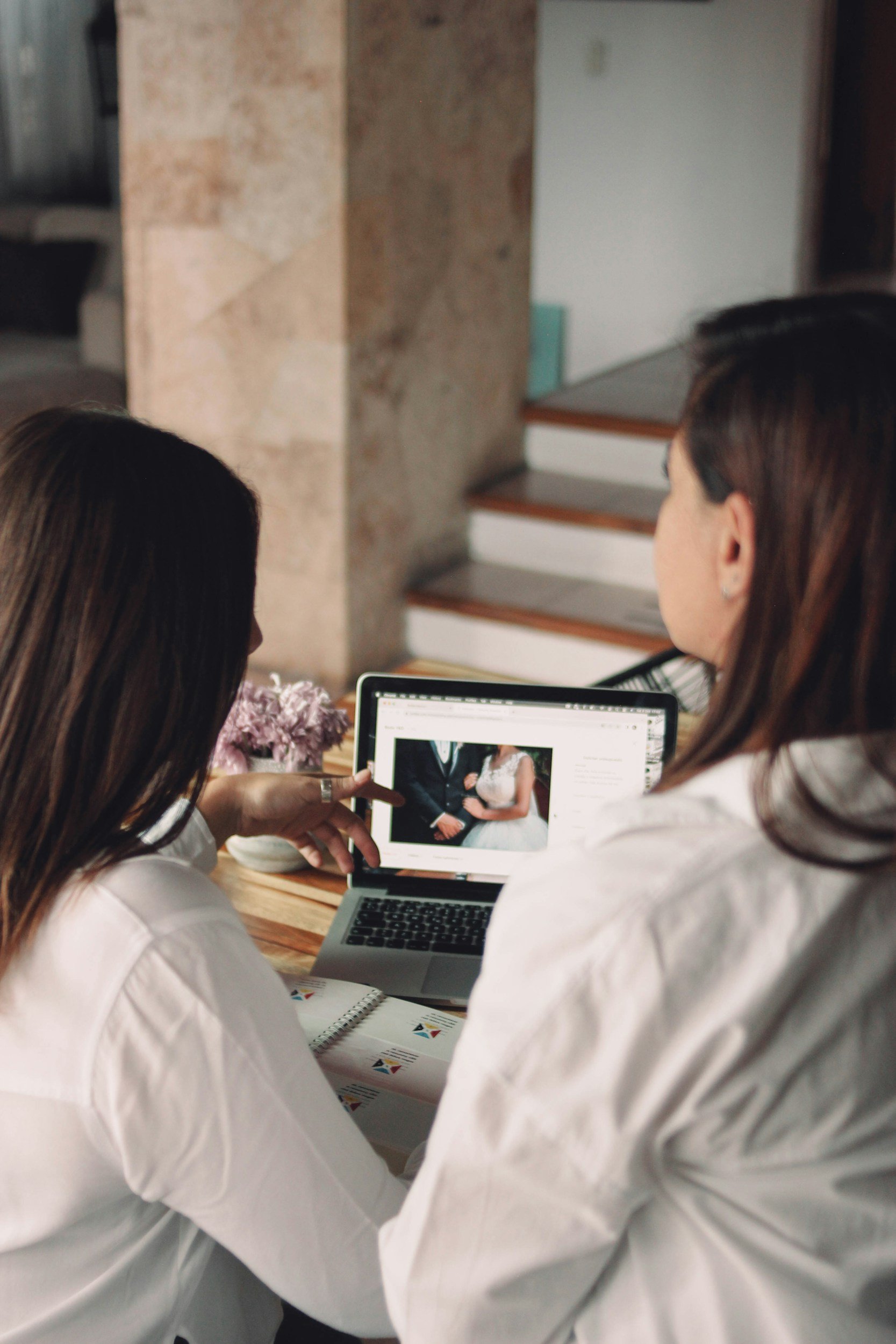 Two women reviewing wedding photo on laptop in a cozy indoor setting.
