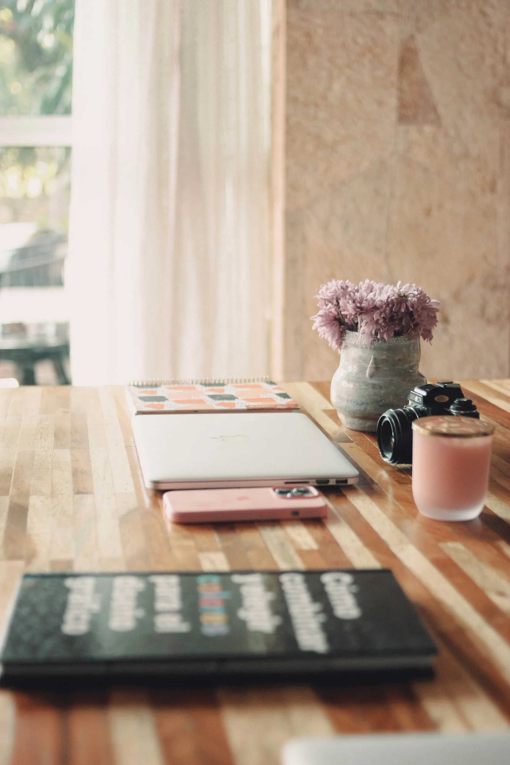 Wooden table with a laptop, notebook, pink phone, camera, pink candle holder, and a vase with pink flowers, set against a window with curtains.