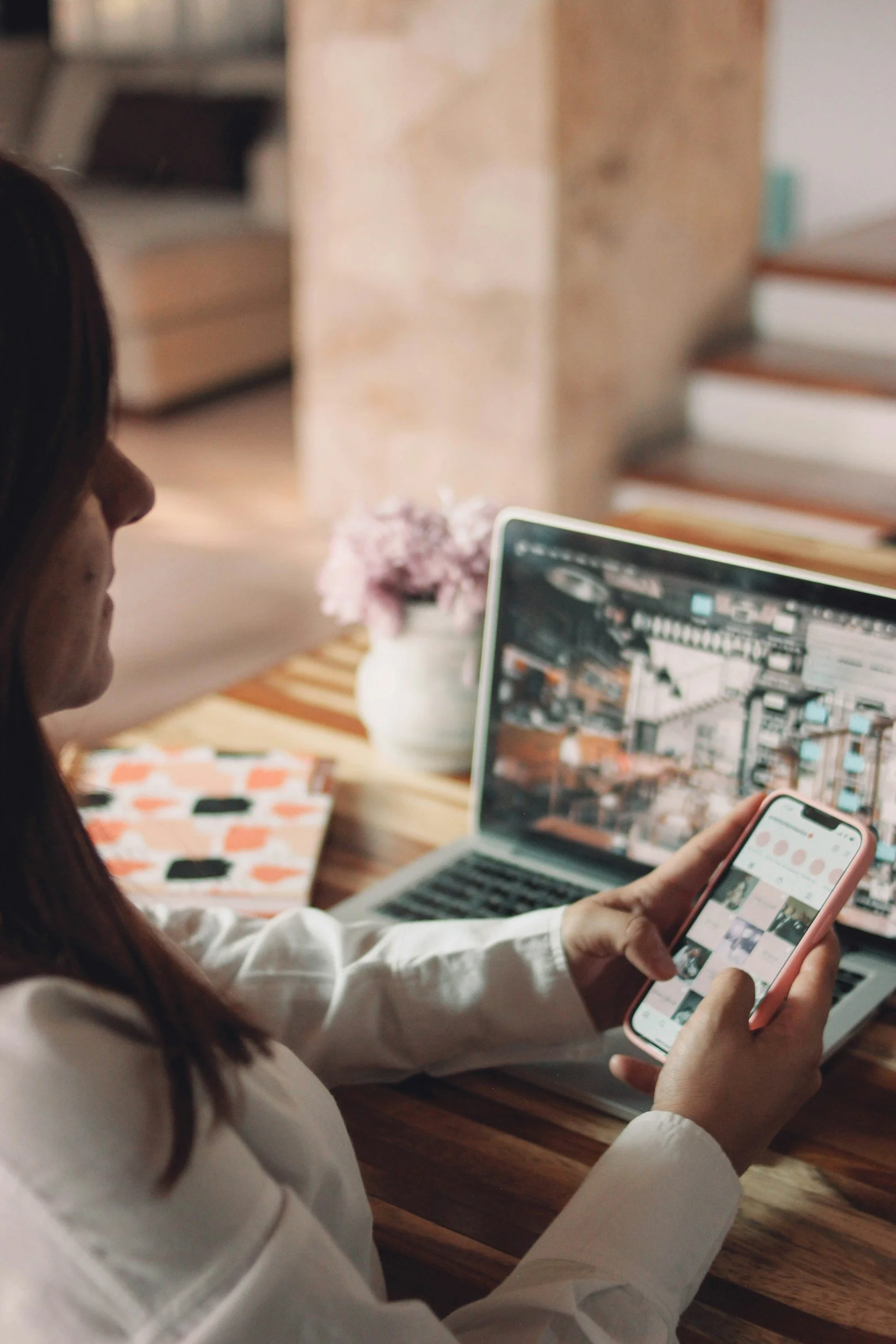 A woman sitting at a wooden table, using a smartphone, with a laptop open in front of her displaying an indoor scene, and a pink notebook beside the laptop. A vase with pink flowers is in the background.