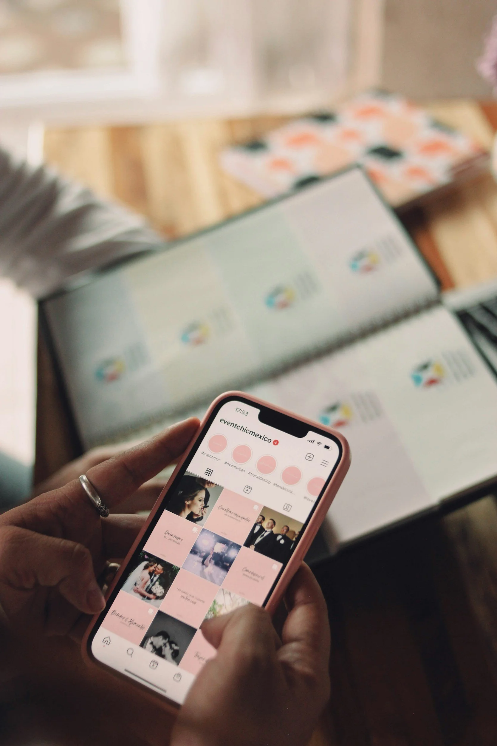 Person holding a smartphone displaying an Instagram profile called 'eventchimexico,' with photos of people and events, against a background of a table with open documents or photo albums.