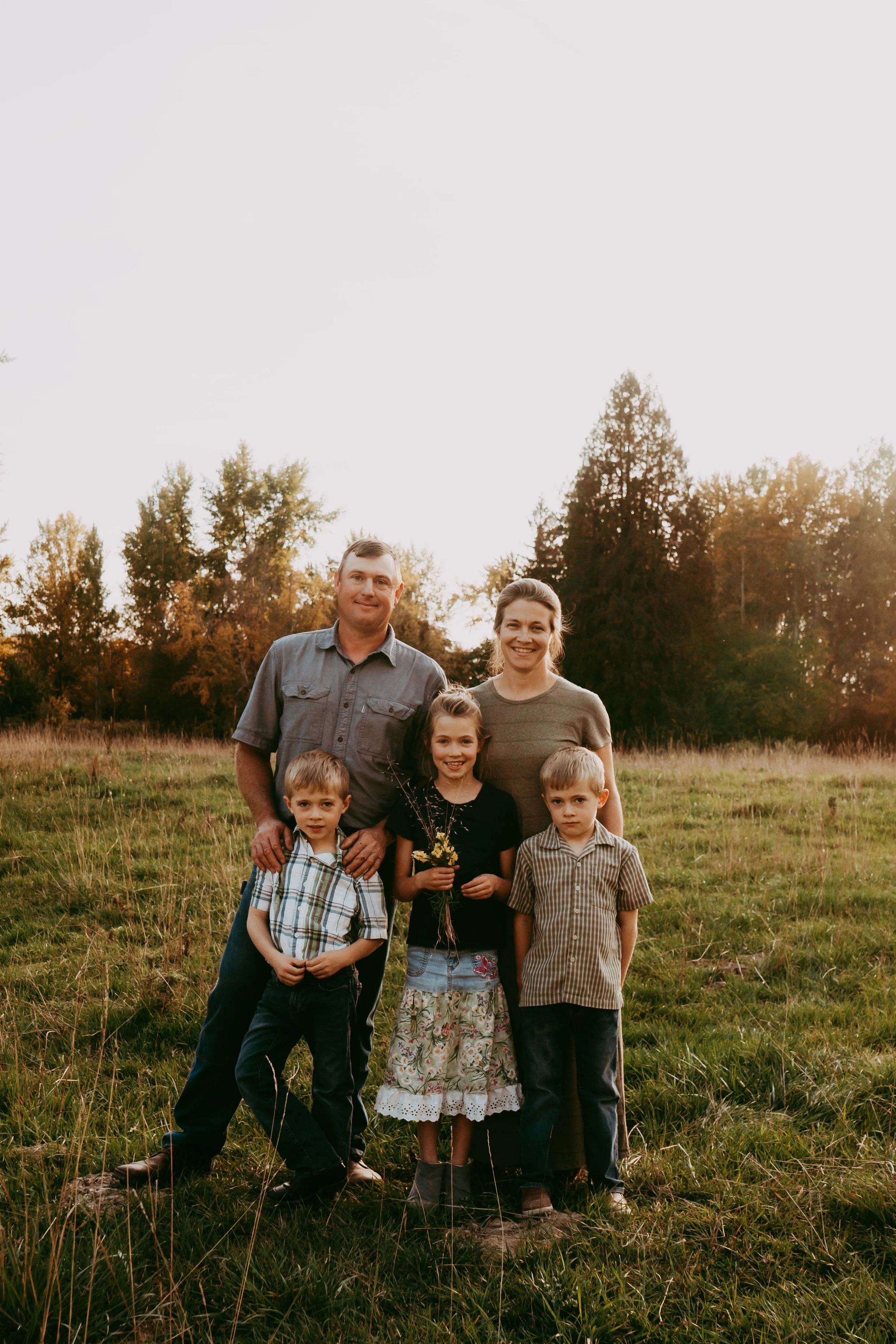 Family of five farmers standing outdoors in a grassy field during sunset, smiling at the camera.