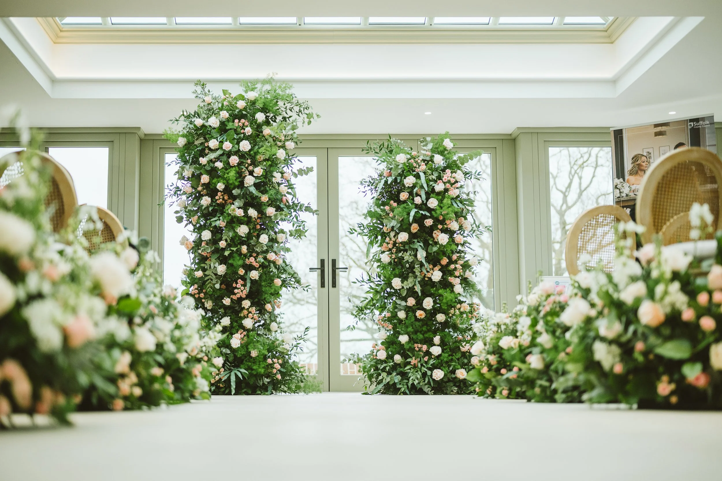 Elegant indoor space decorated with tall floral arrangements of roses, greenery, and other flowers in pastel colors, with glass doors and large windows.