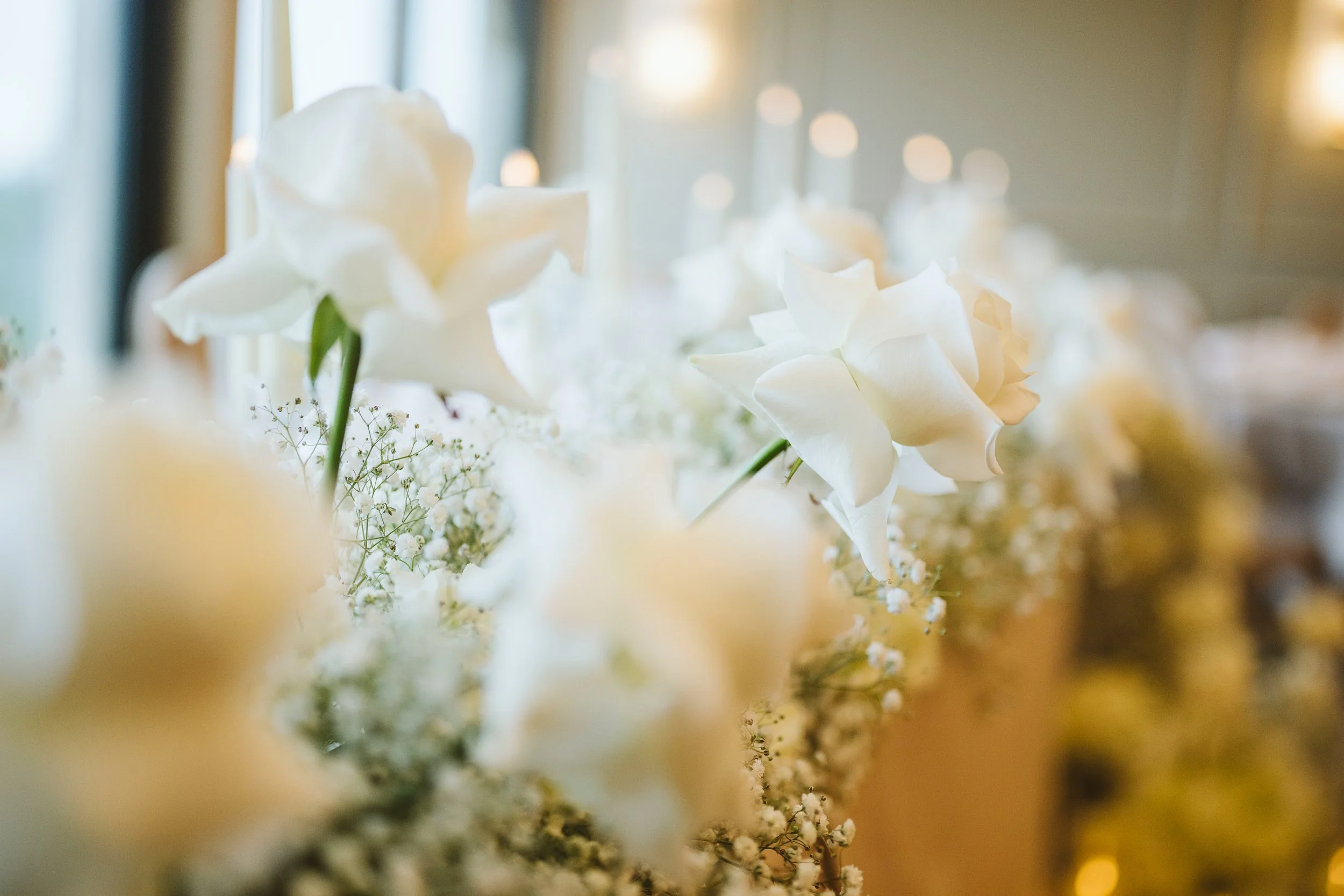 Close-up of white flowers, including roses and baby's breath, on a blurred background, likely at a wedding or event reception.