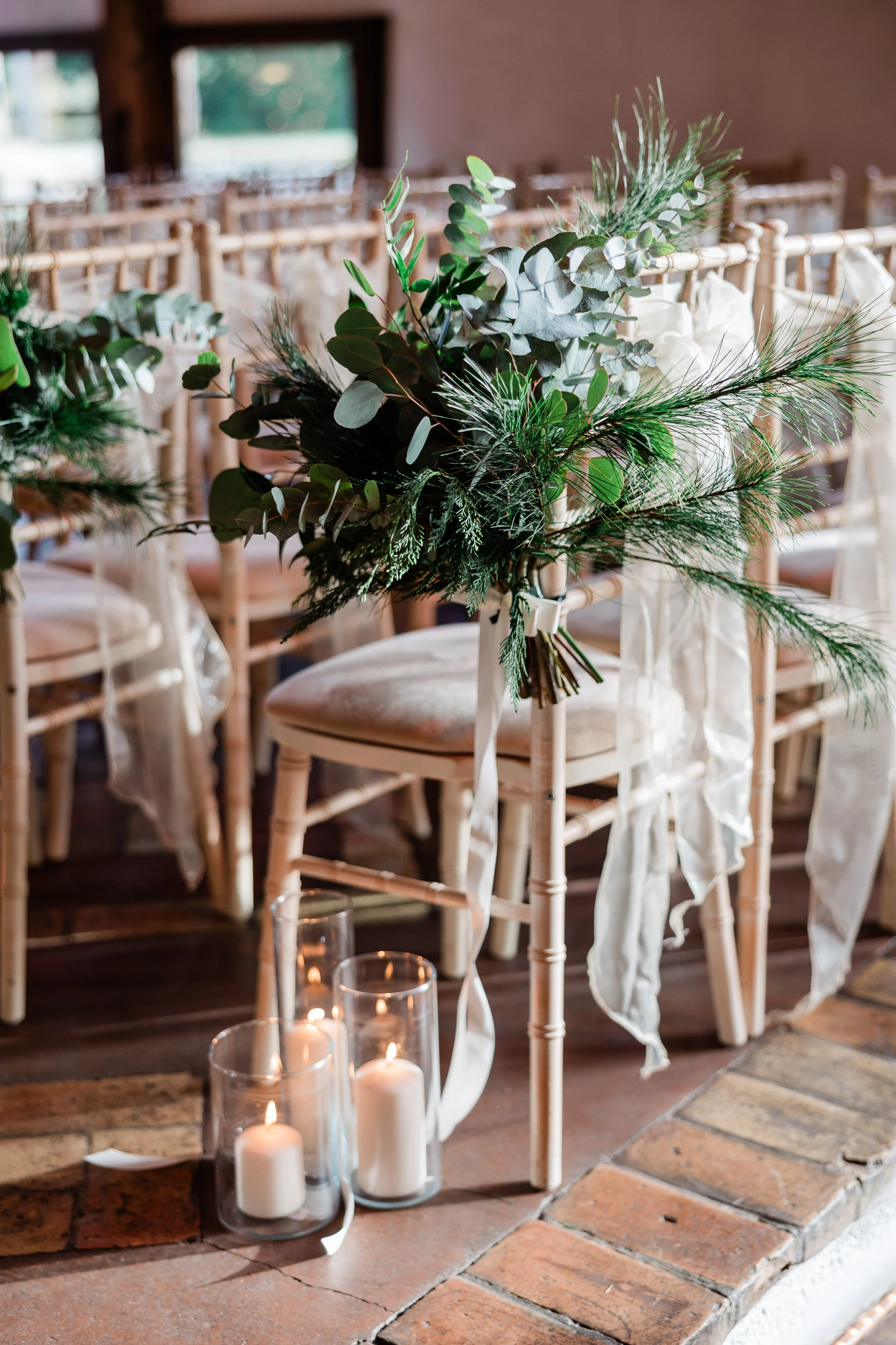 Decorative wedding or event chair with a green and white floral arrangement tied to it, placed on a brick floor near candles in glass holders.