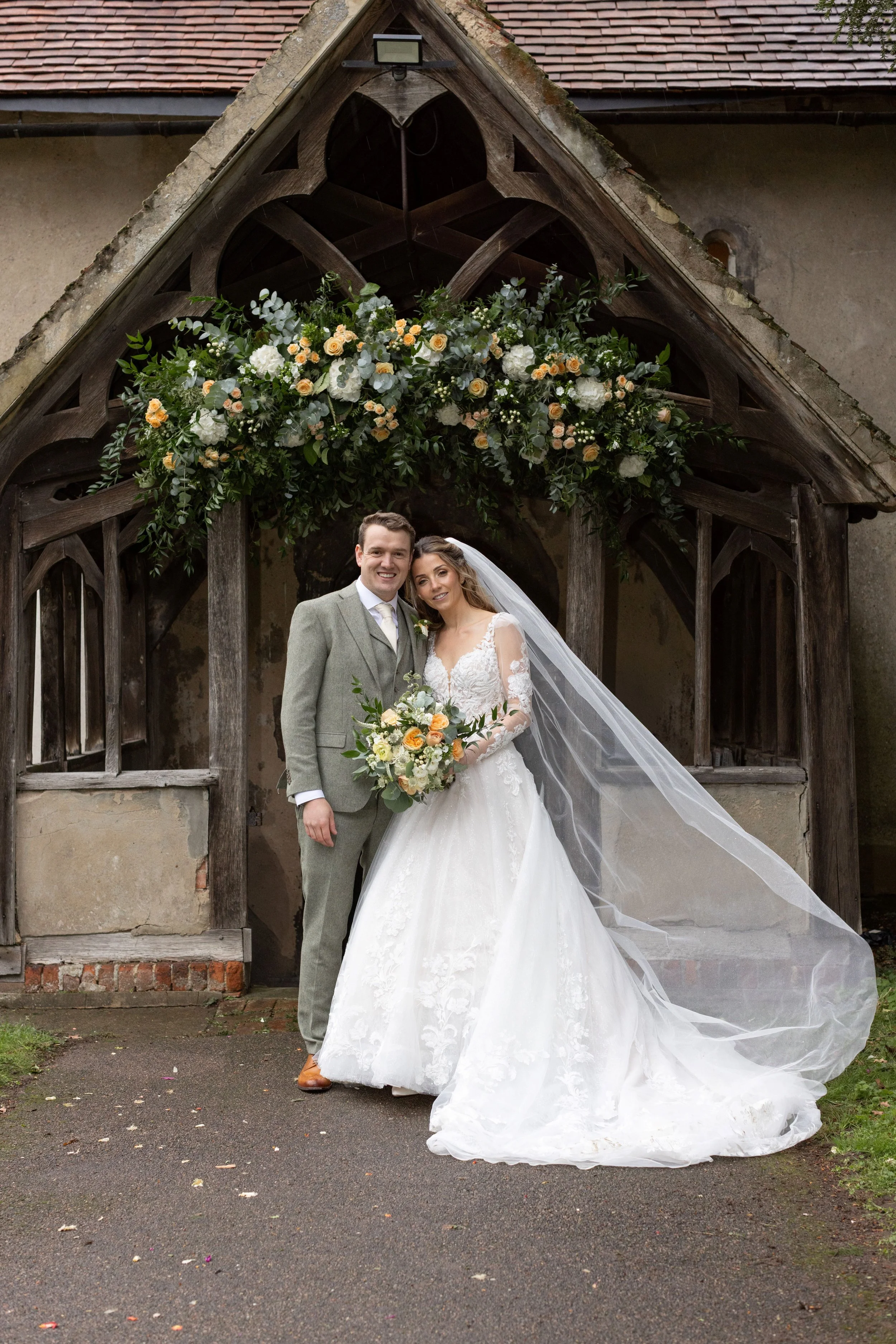 A newlywed couple standing in front of a wooden arch decorated with flowers, the bride in a white lace gown with a long veil holding a bouquet, and the groom in a gray suit.
