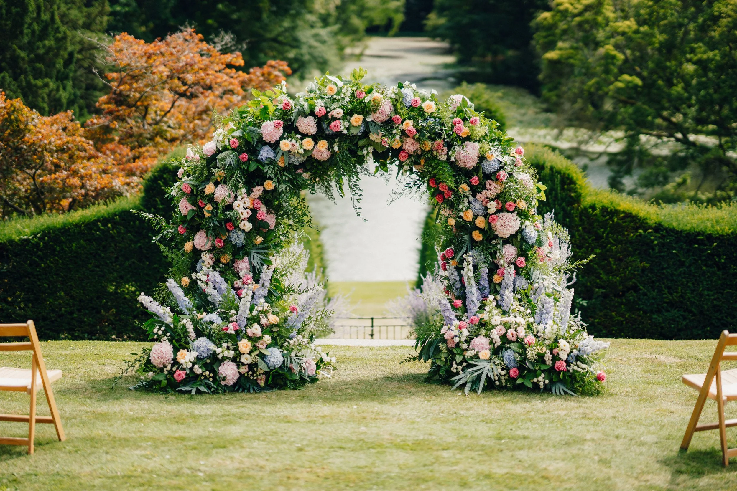 An outdoor wedding arch decorated with pink, purple, and white flowers, set in a lush green garden near a river.