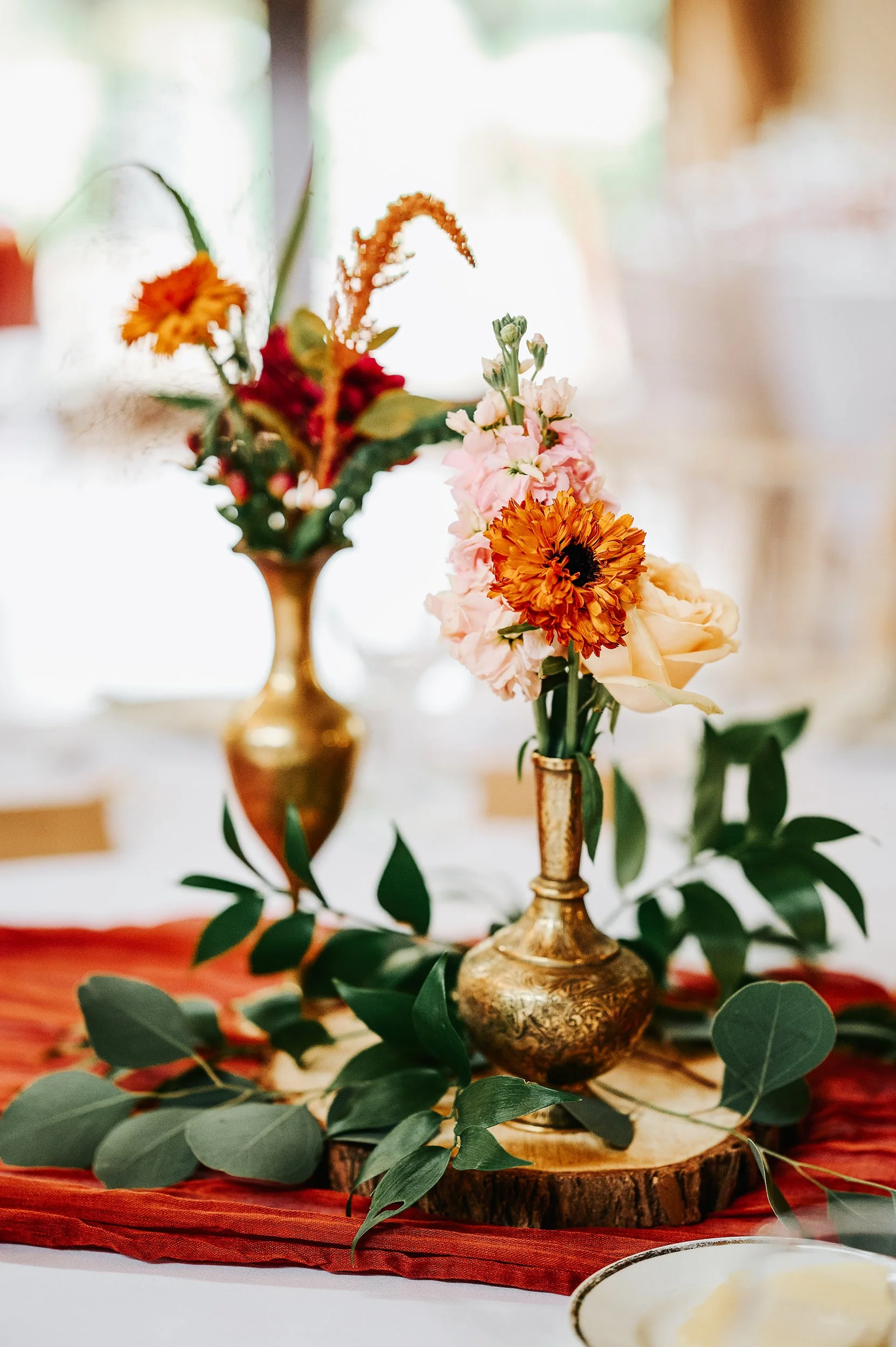 Elegant floral arrangement in gold vases with pink and orange flowers, greenery, and a wood slice base, on a red table runner.