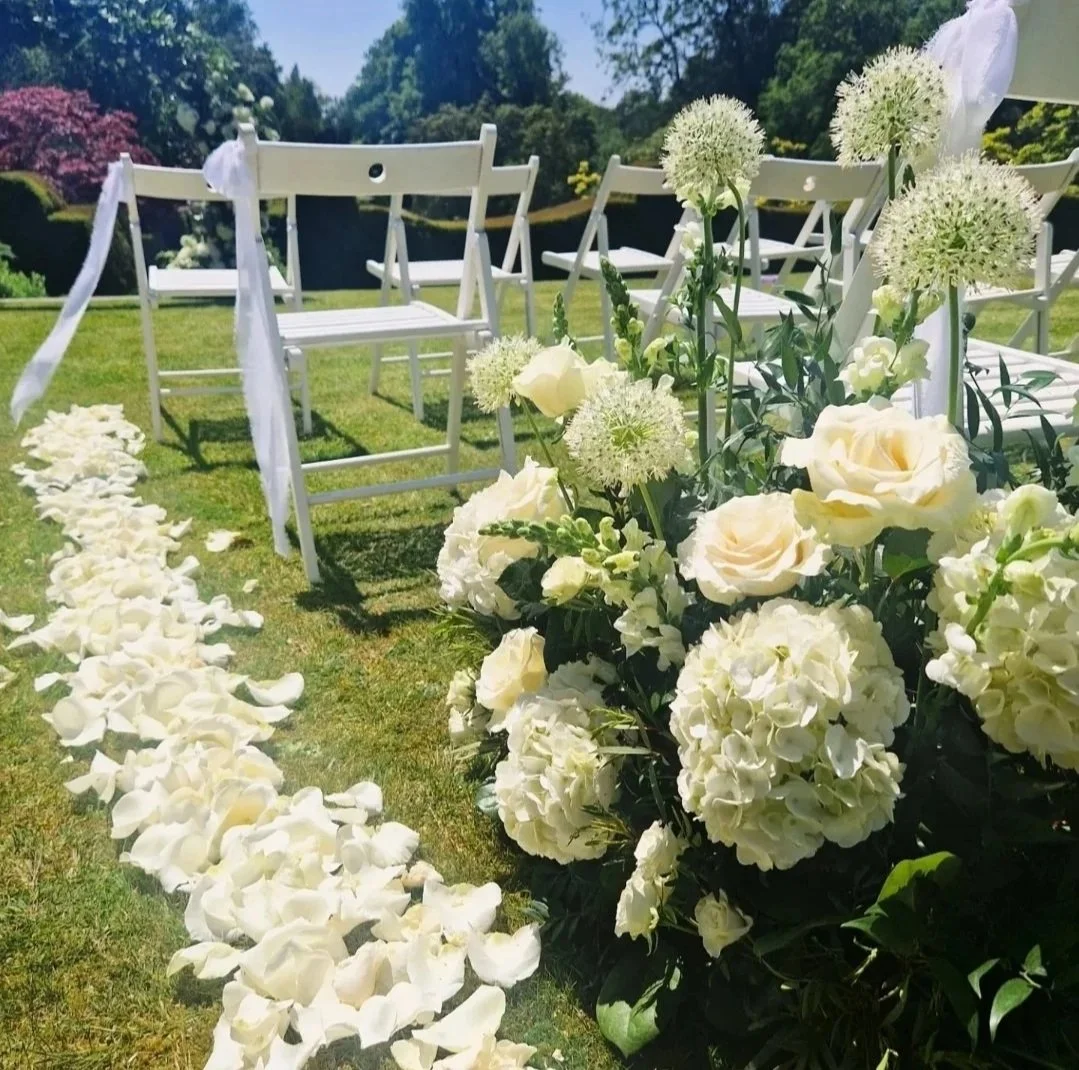Outdoor wedding setup with white wooden chairs decorated with white ribbons, flower petals on the grass aisle, and large white flowers in the foreground, set in a lush garden with trees and shrubs in the background.