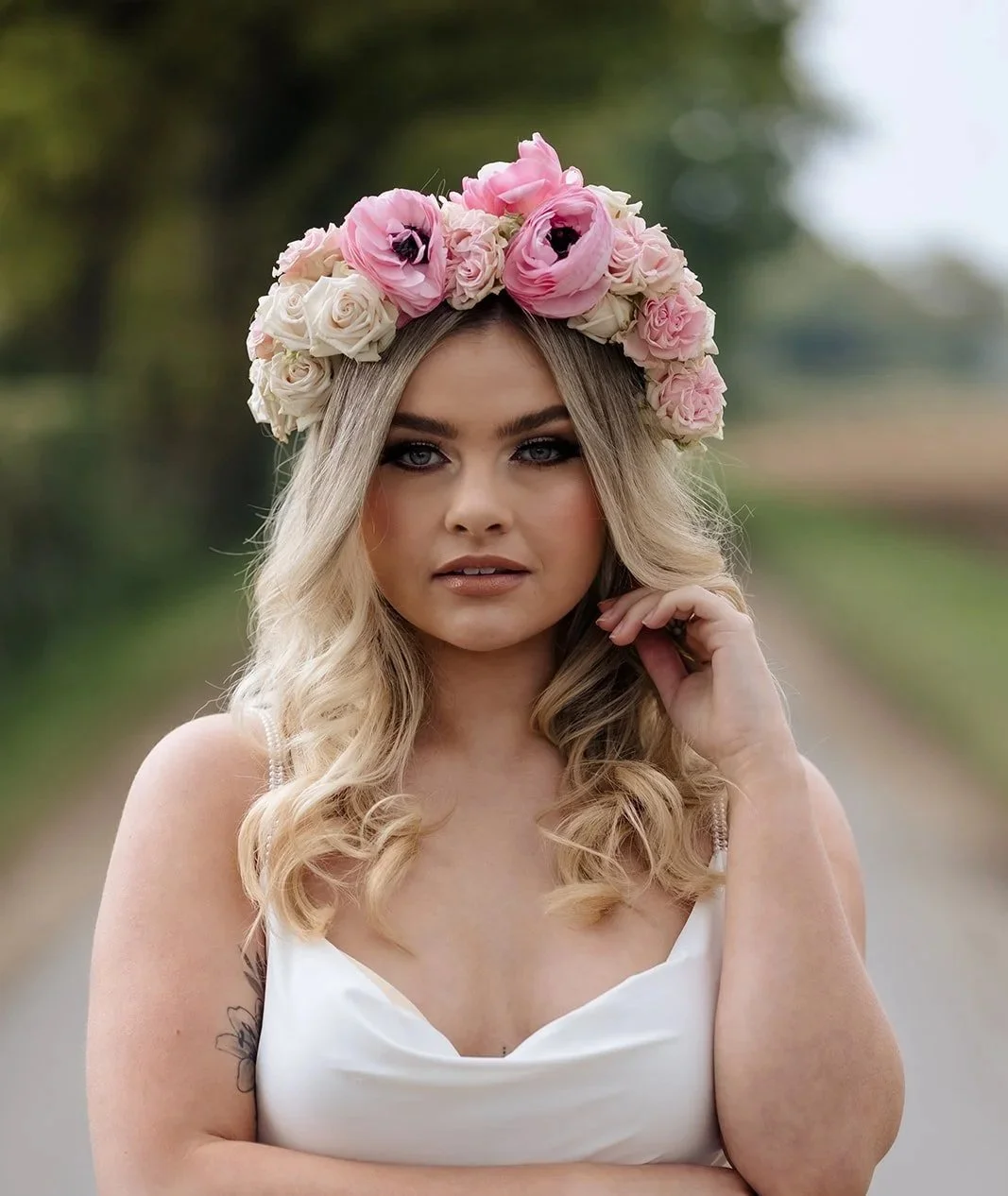 A woman with blonde hair wearing a floral crown of pink and white roses and black anemone flowers, and a white sleeveless top. She is standing outdoors on a blurred natural background, with her hand near her face.