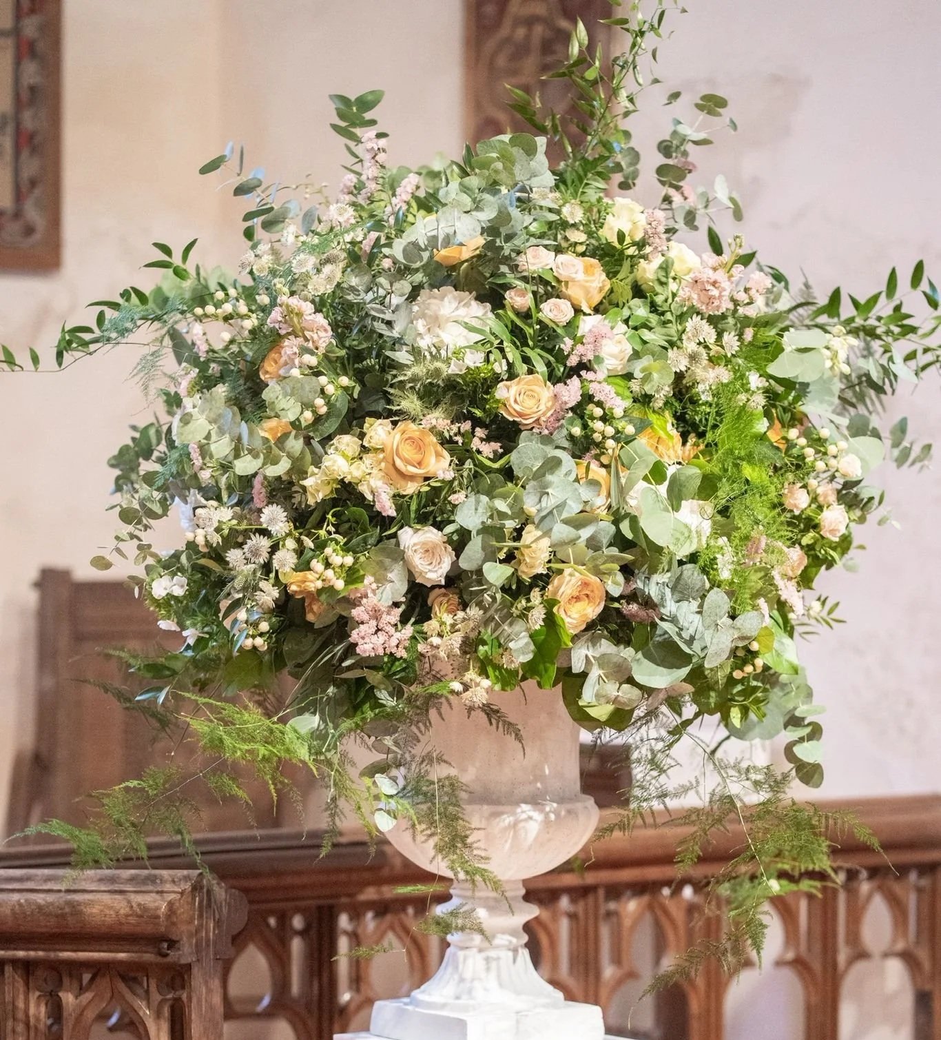 Large floral arrangement with cream and pink roses, white and pink blossoms, and green foliage in a white pedestal urn on a wooden surface.