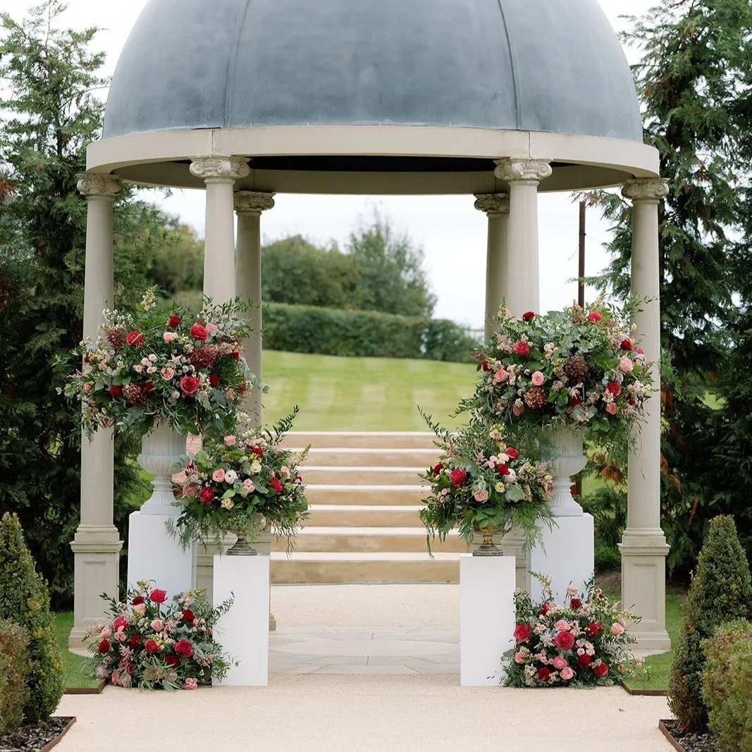 An outdoor wedding or ceremony setup featuring a small white gazebo with a domed roof, decorated with large floral arrangements in urns and on white pedestals in shades of pink, red, and white, with lush green trees and a grassy lawn in the backgroun