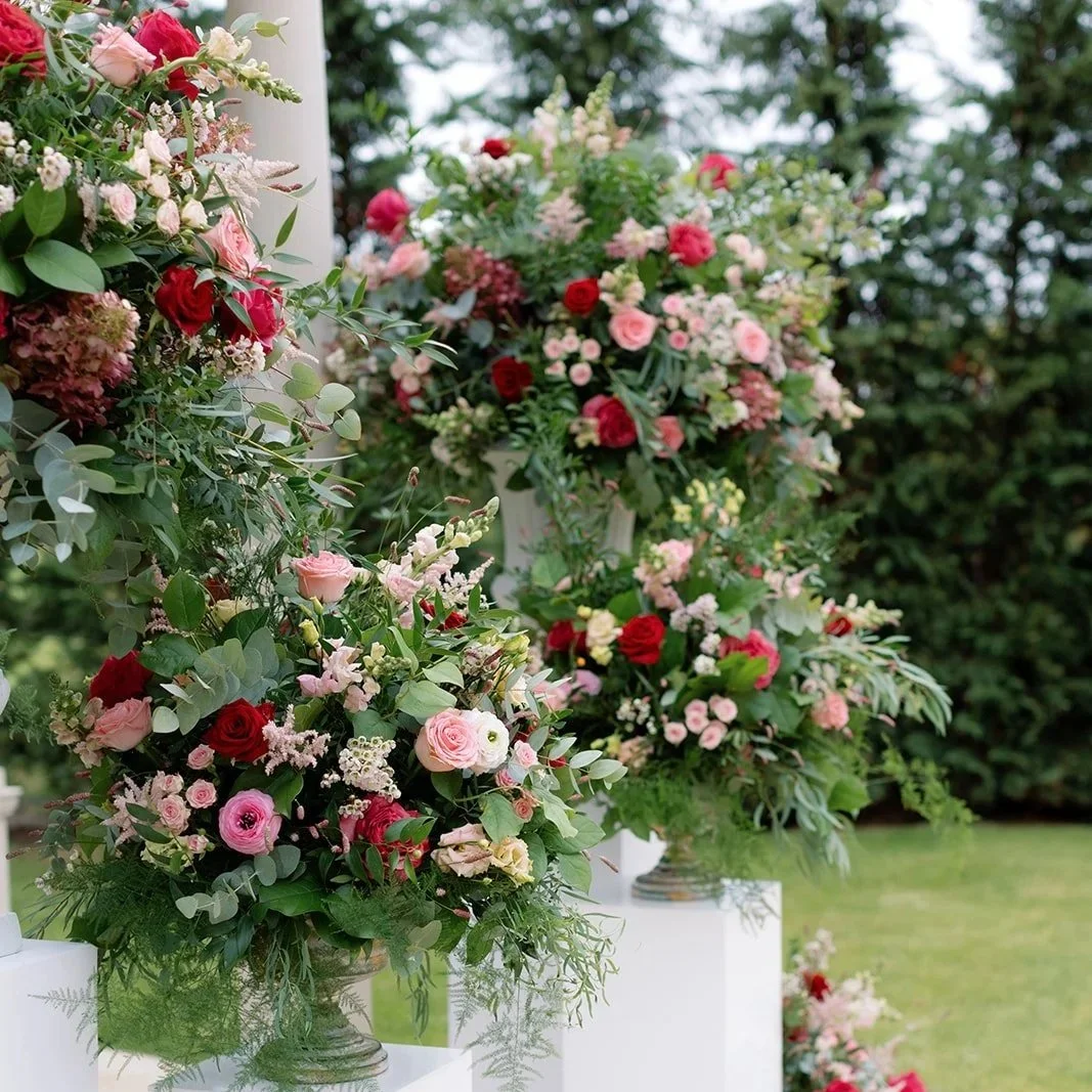 Array of floral arrangements with pink, red, and white roses on white pedestals outside, with green trees and grass in the background.