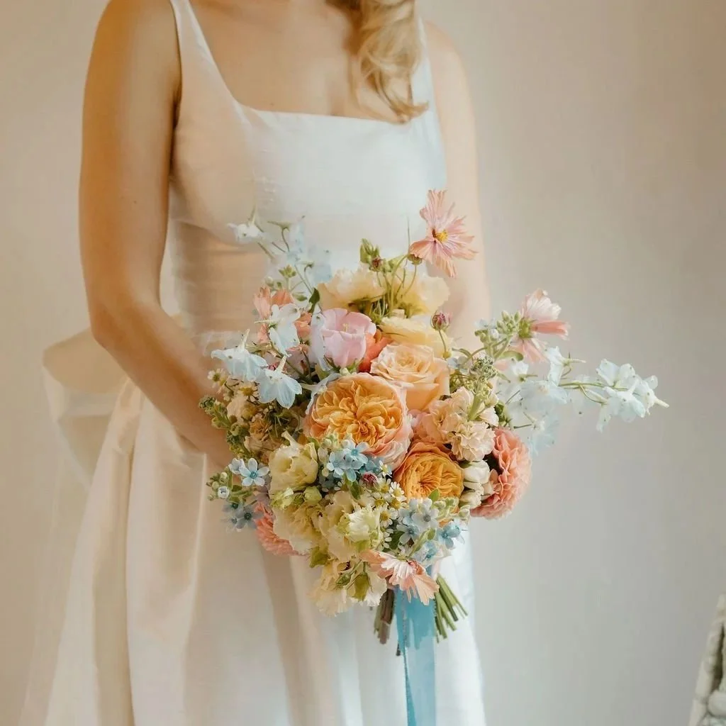 Woman in a satin wedding dress holding a large bouquet of pastel-colored flowers, including roses, delphiniums, and other blossoms, accented with a blue ribbon.