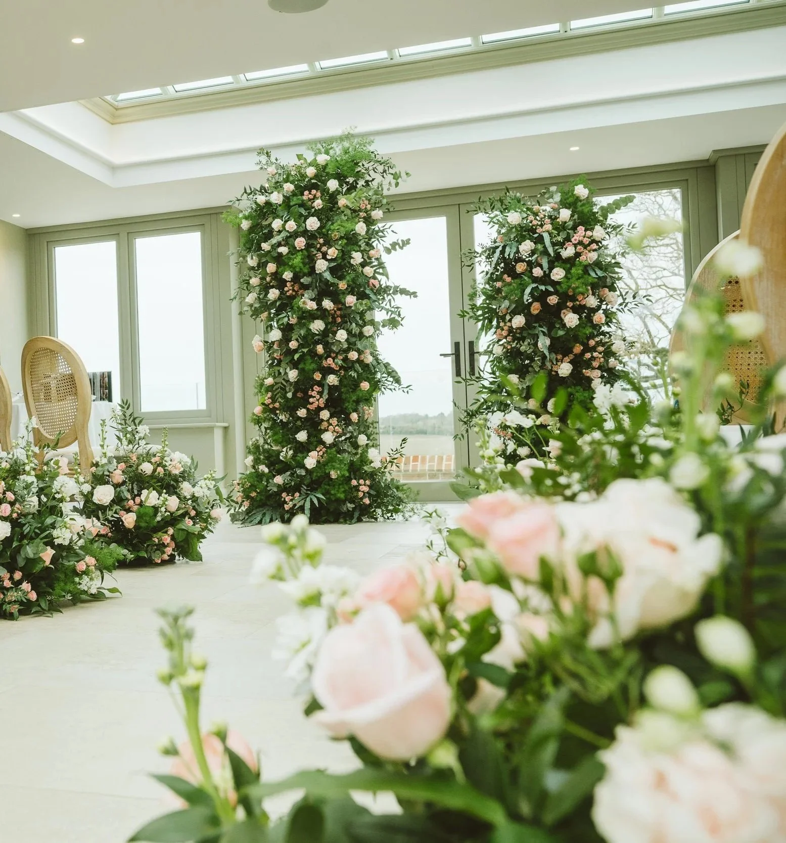 Elegant indoor wedding ceremony setup with floral arches and arrangements of pink and white roses and greenery, natural light streaming through large windows.