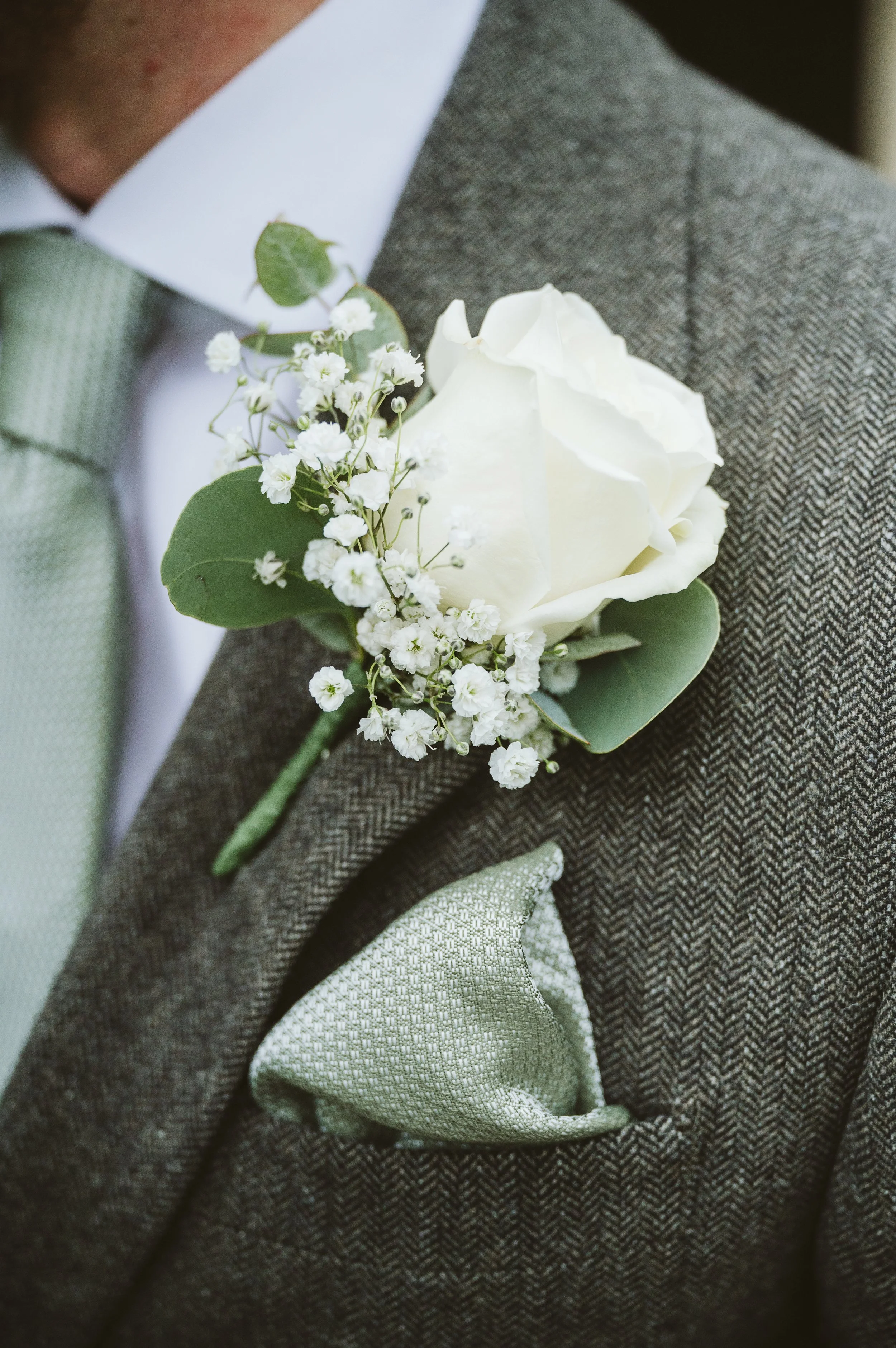 Close-up of a men's boutonniere with a white rose, baby's breath, and eucalyptus leaves pinned to a brown tweed jacket, with a light green pocket square in the breast pocket.
