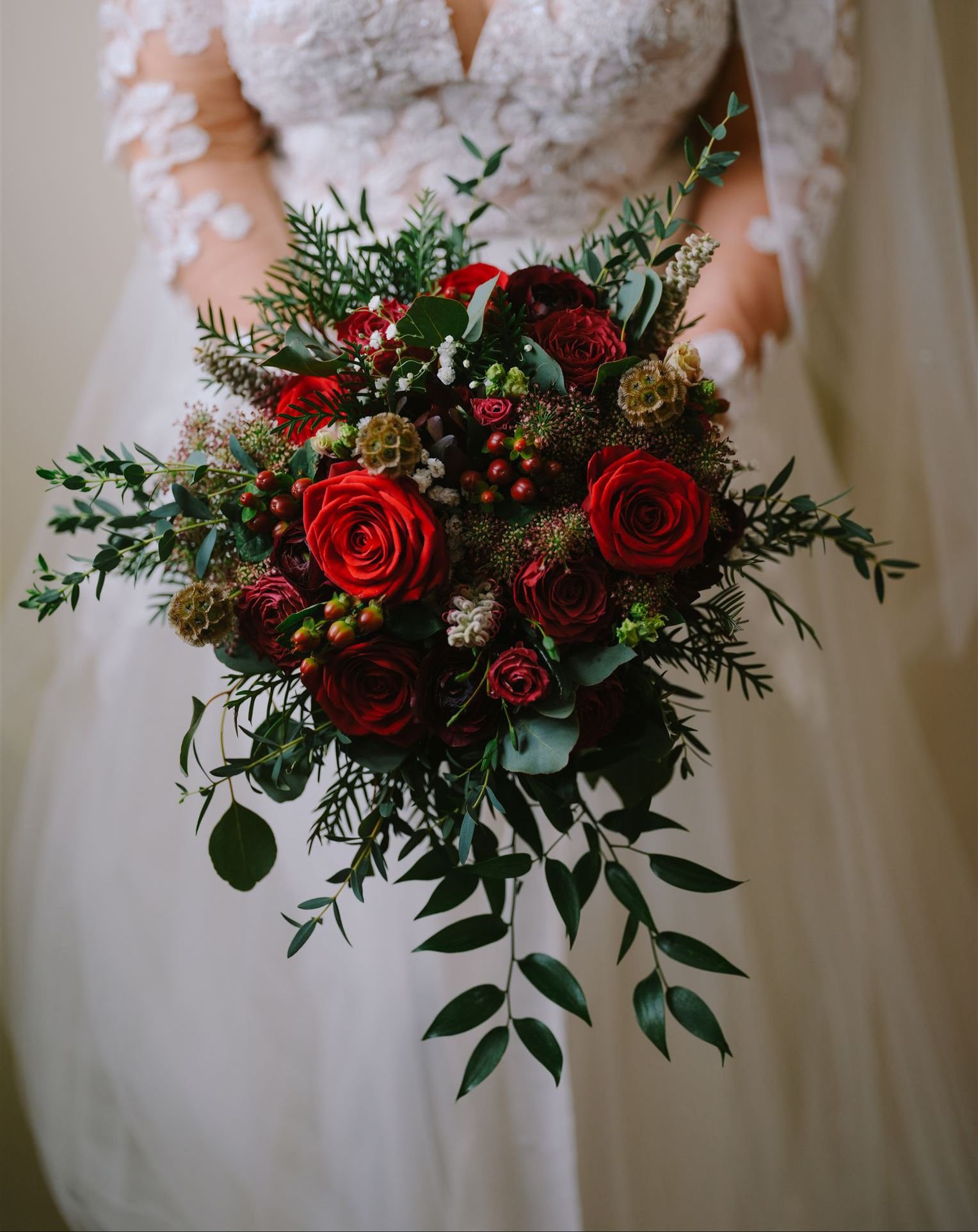 Bride holding a lush wedding bouquet with red roses, greenery, and small white flowers.