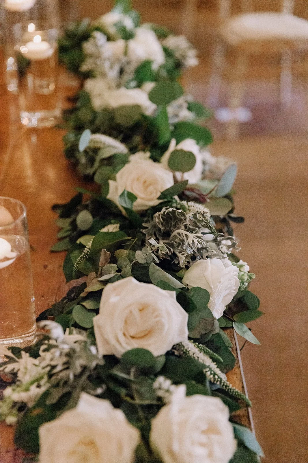 A floral centerpiece with white roses and greenery on a wooden table, decorated with candles and glasses in a cozy, warmly lit setting.