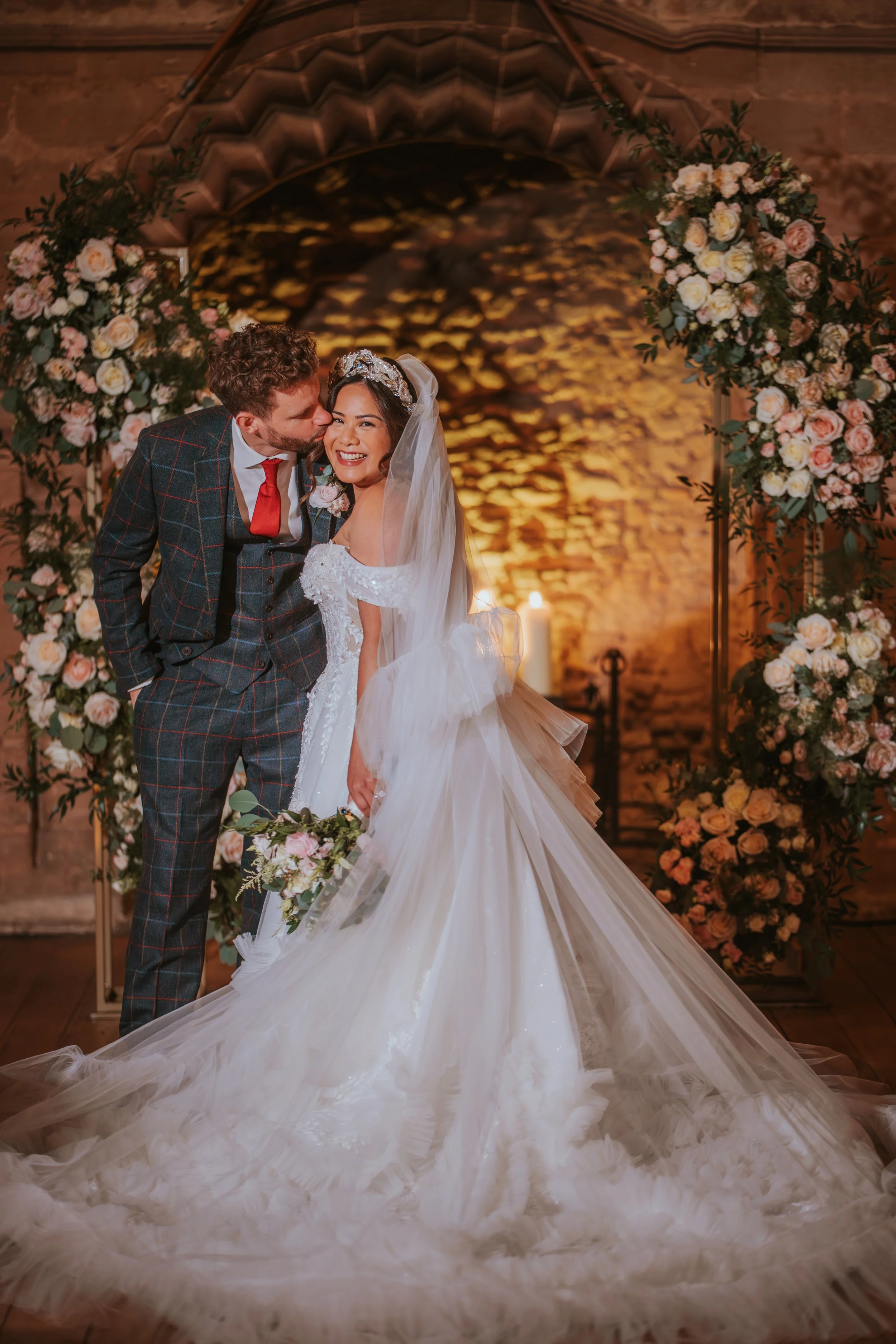 A bride and groom standing under a floral arch, smiling and sharing a moment on their wedding day. The bride is wearing a white wedding gown with a long train and veil, holding a bouquet of flowers, while the groom is dressed in a checkered suit with