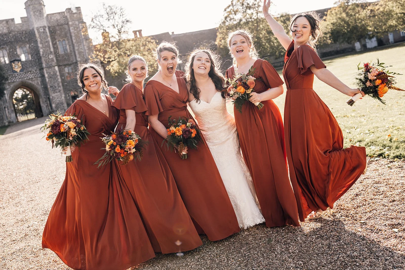 A group of six women in matching rust-colored bridesmaid dresses and a bride in a white wedding gown, standing outside near a historic stone castle, celebrating and smiling, holding bouquets of flowers.